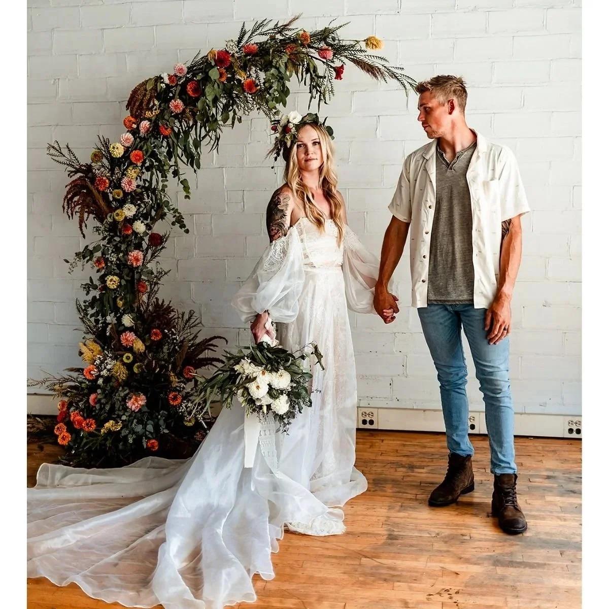 A bride and groom standing under a floral arch, holding hands, with a white brick wall in the background. The bride is wearing a white wedding dress with puffed sleeves and a floral headpiece, holding a bouquet. The groom is dressed in casual attire with a short-sleeved shirt, T-shirt, jeans, and boots.