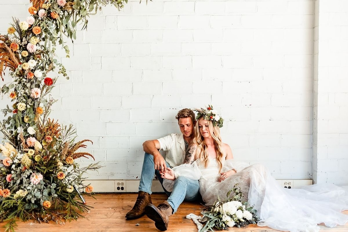 A couple sits on the wooden floor in front of a white brick wall, dressed in wedding attire. The woman wears a white wedding dress and a flower crown, while the man wears a light-colored shirt, jeans, and brown boots. They are holding hands and sitting close together near a wedding bouquet. A large floral arch decorated with various flowers and greenery stands to the left.