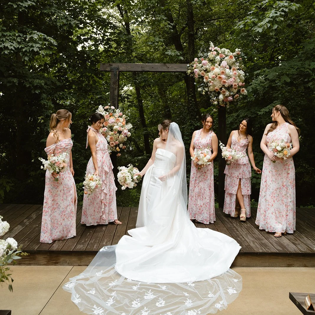 A bride in a white wedding dress and veil holding a bouquet stands on a wooden platform, surrounded by five bridesmaids in pink floral dresses holding bouquets, at an outdoor wedding ceremony with a floral arch and green trees in the background.