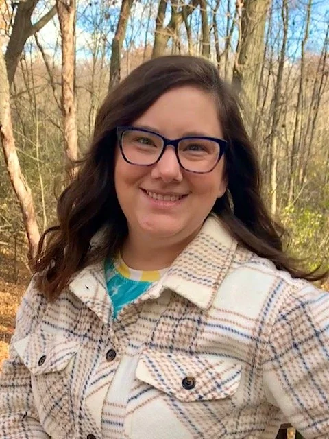 A smiling woman with glasses and long brown hair stands outdoors in a wooded area during autumn.