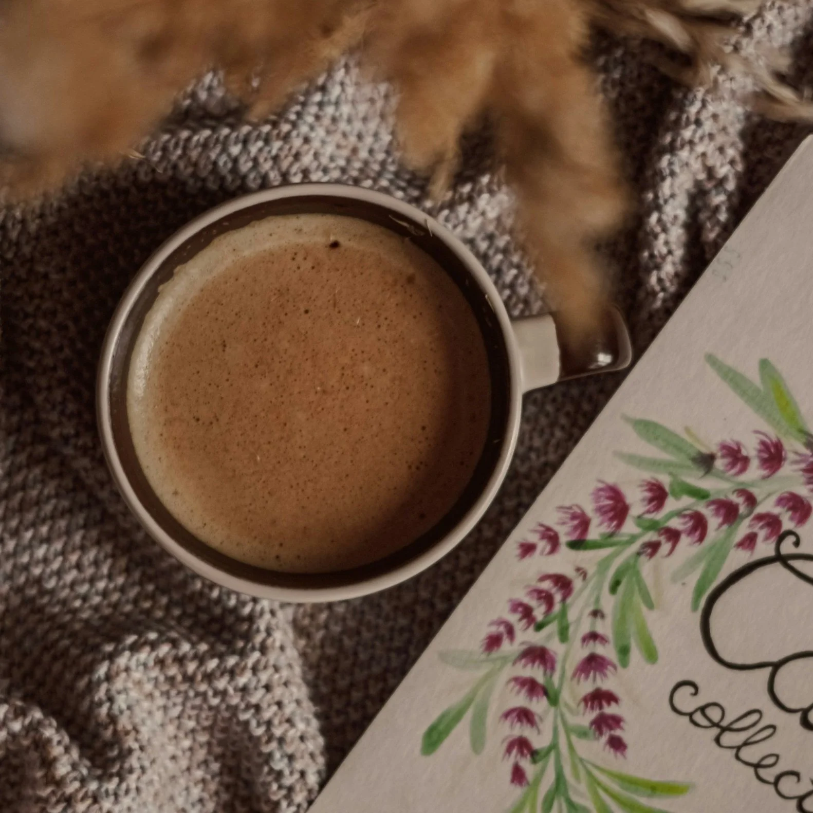 A cup of coffee on a textured knit blanket with a decorative floral design on a piece of paper nearby.