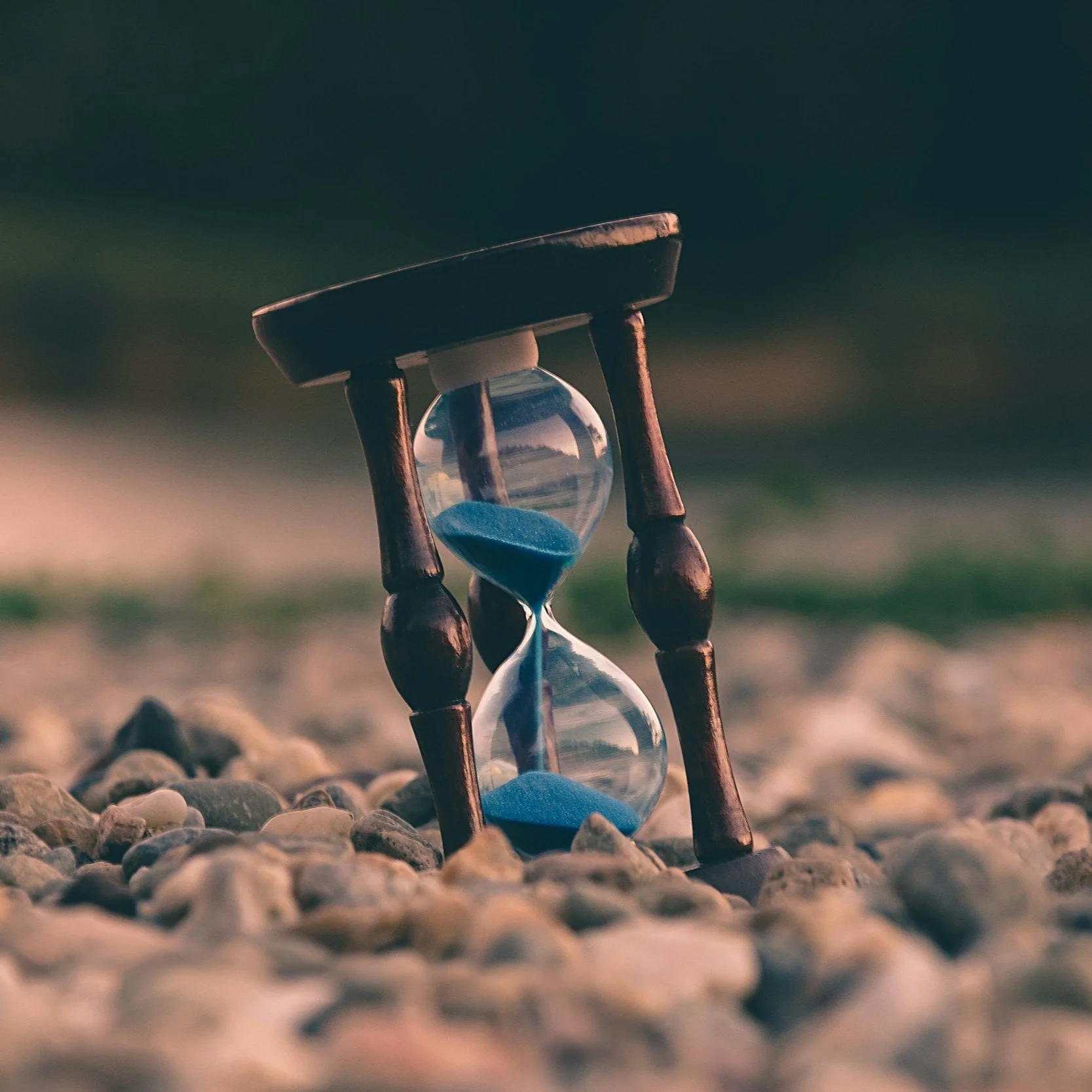 A wooden hourglass with blue sand on pebbles outdoors.