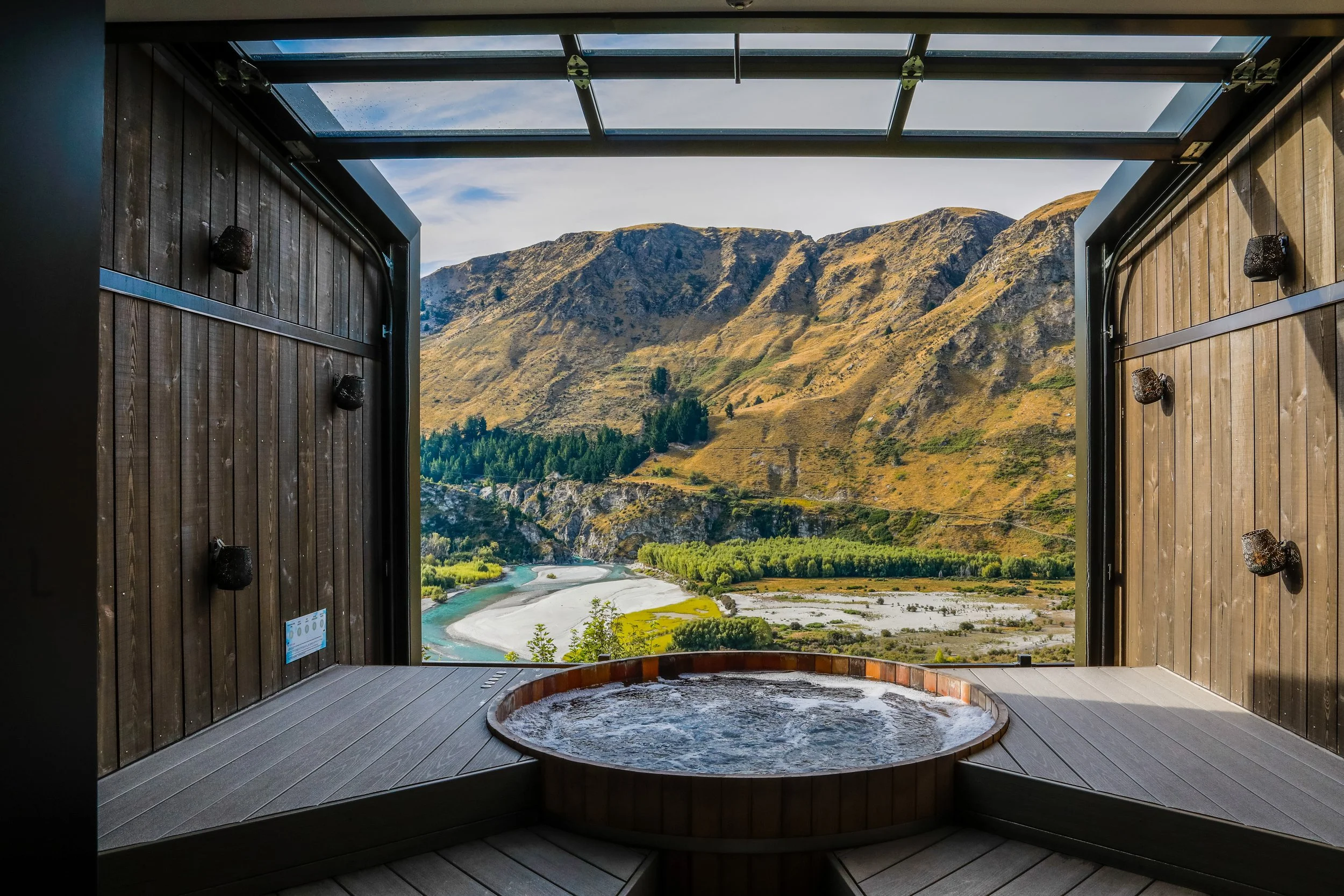 Wooden outdoor hot tub on a deck overlooking mountains, river, and green scenery.