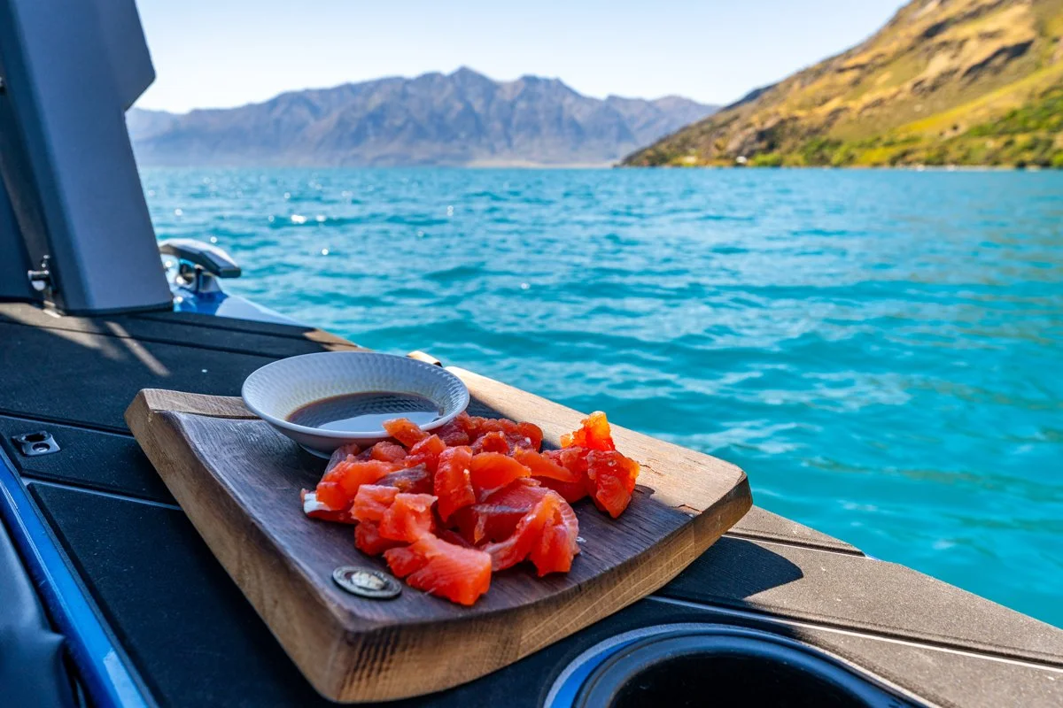 Sliced salmon sashimi with soy sauce on a wooden board, placed on a boat with a blue water lake and mountains in the background.