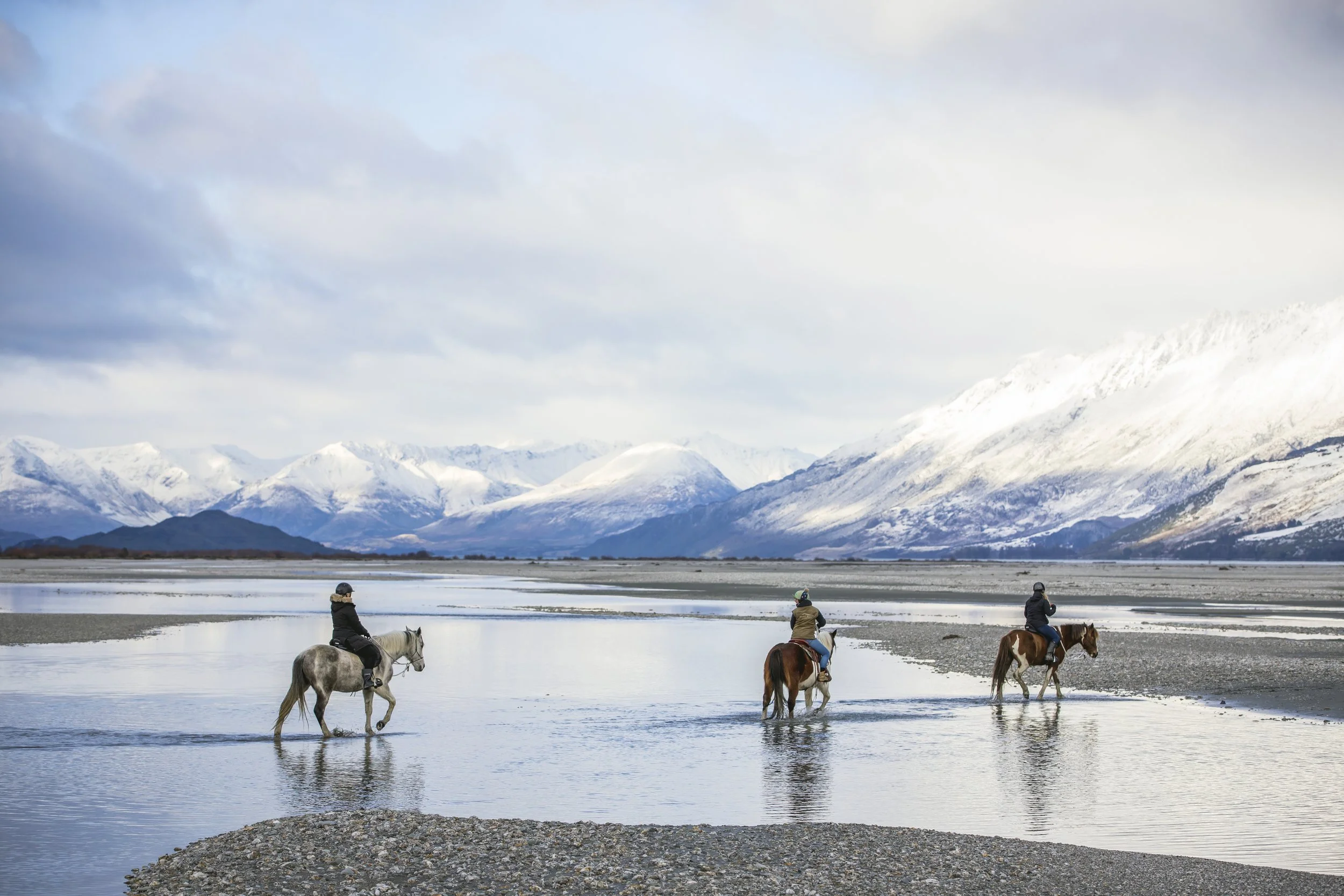 Three people riding horses through shallow water with snow-capped mountains in the background.