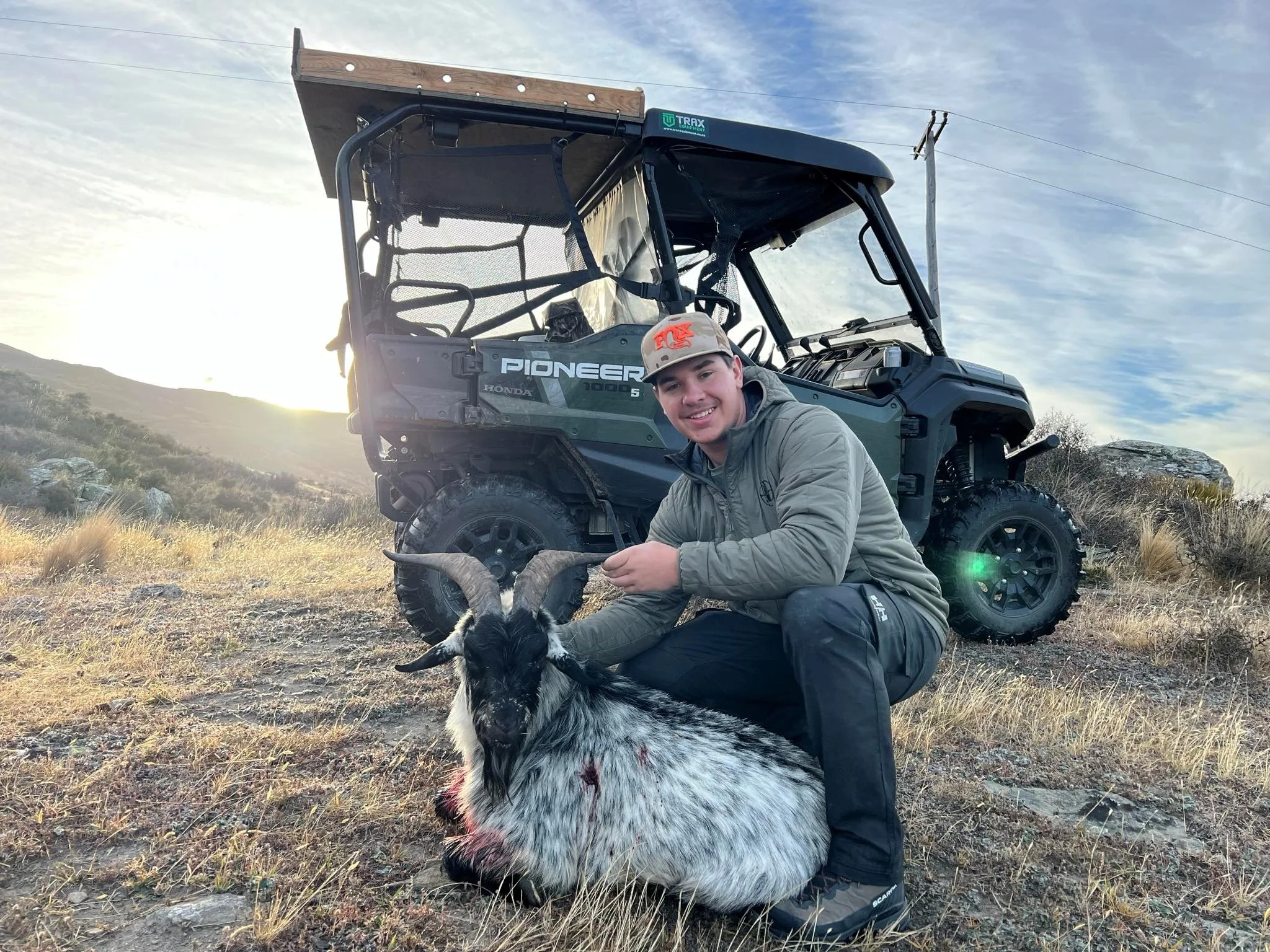 A young man in outdoor gear kneeling next to a dead goat with large curved horns on a grassy and rocky hillside, with a Honda Pioneer ATV in the background and the sun setting behind distant hills.
