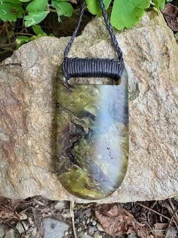 A handcrafted pendant with green, black, and brown swirl design, hanging from a black cord, resting on a large rock.