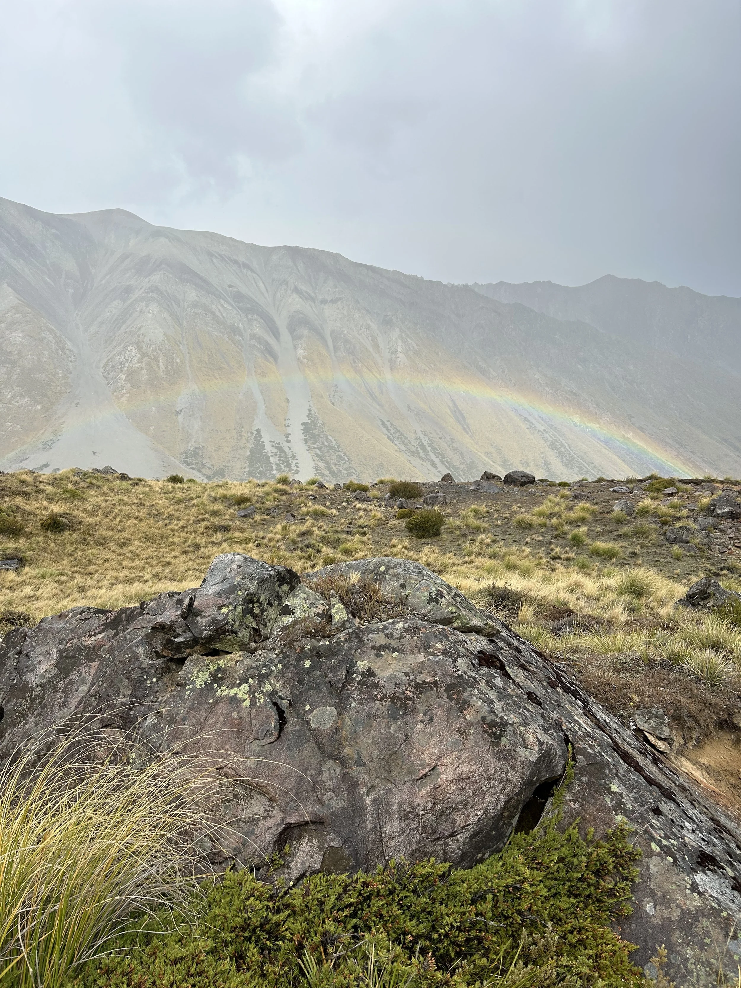 A mountainous landscape with a rainbow arching across the sky, a rocky foreground with grass, and a cloudy sky.