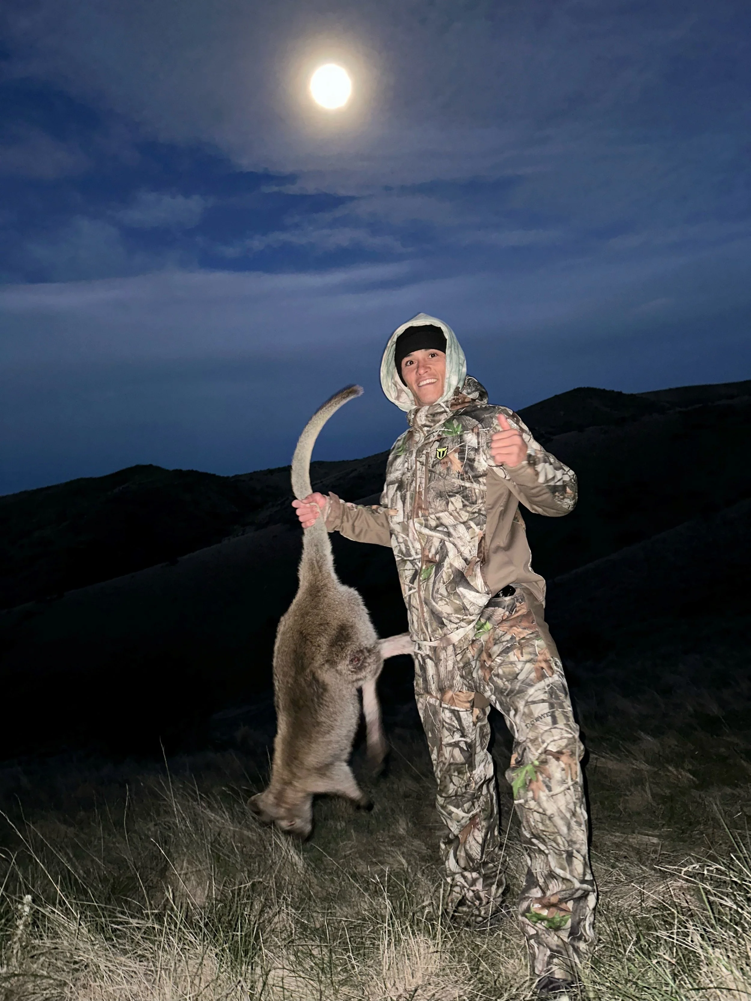 A woman in camouflage outdoor gear holding a dead animal, likely a coyote, under a moonlit sky with rolling hills in the background.