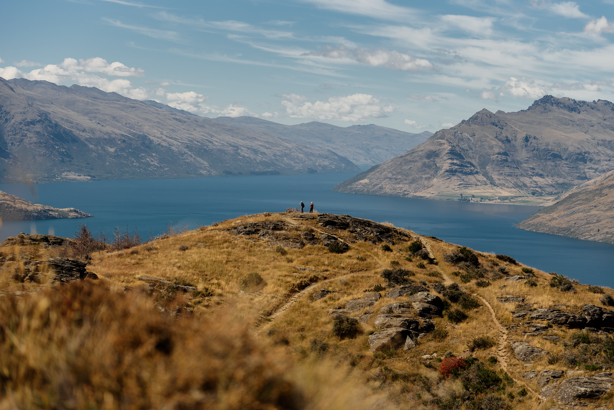Two hikers standing on a hilltop overlooking a large blue lake surrounded by mountains under a partly cloudy sky.