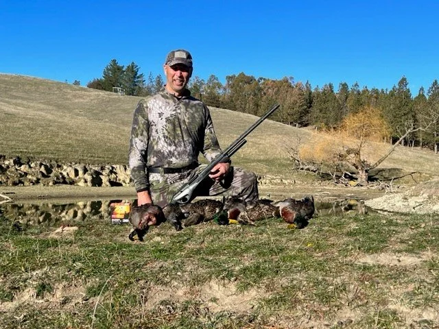 Man with camouflage clothing and sunglasses smiling while holding a rifle, kneeling behind four hunted ducks on grass near a pond with trees in the background.