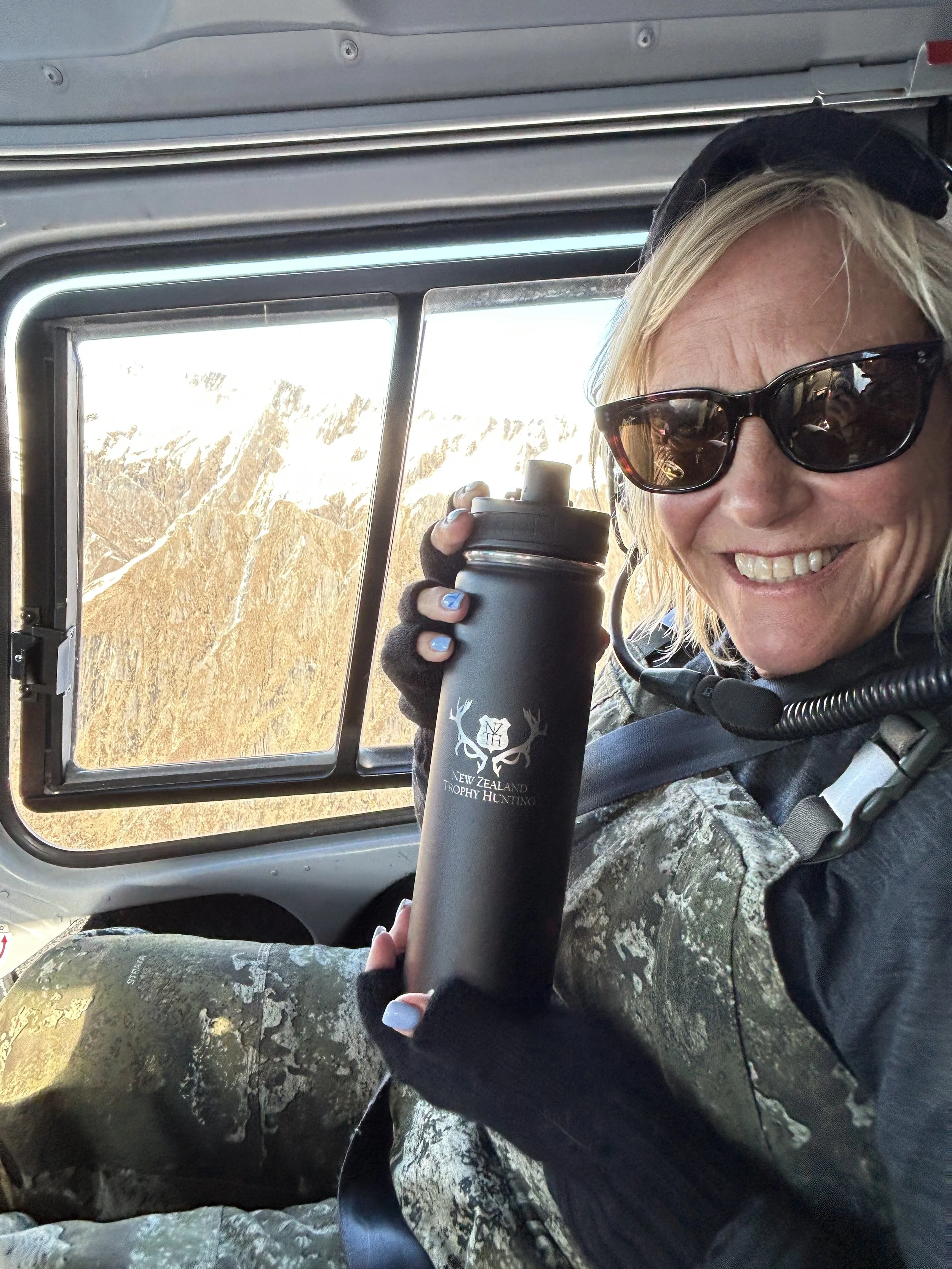 A woman with blonde hair wearing sunglasses and camouflage clothing, smiling while holding a black water bottle with the New Zealand Trophy Hunting logo inside an aircraft with mountains outside the window.