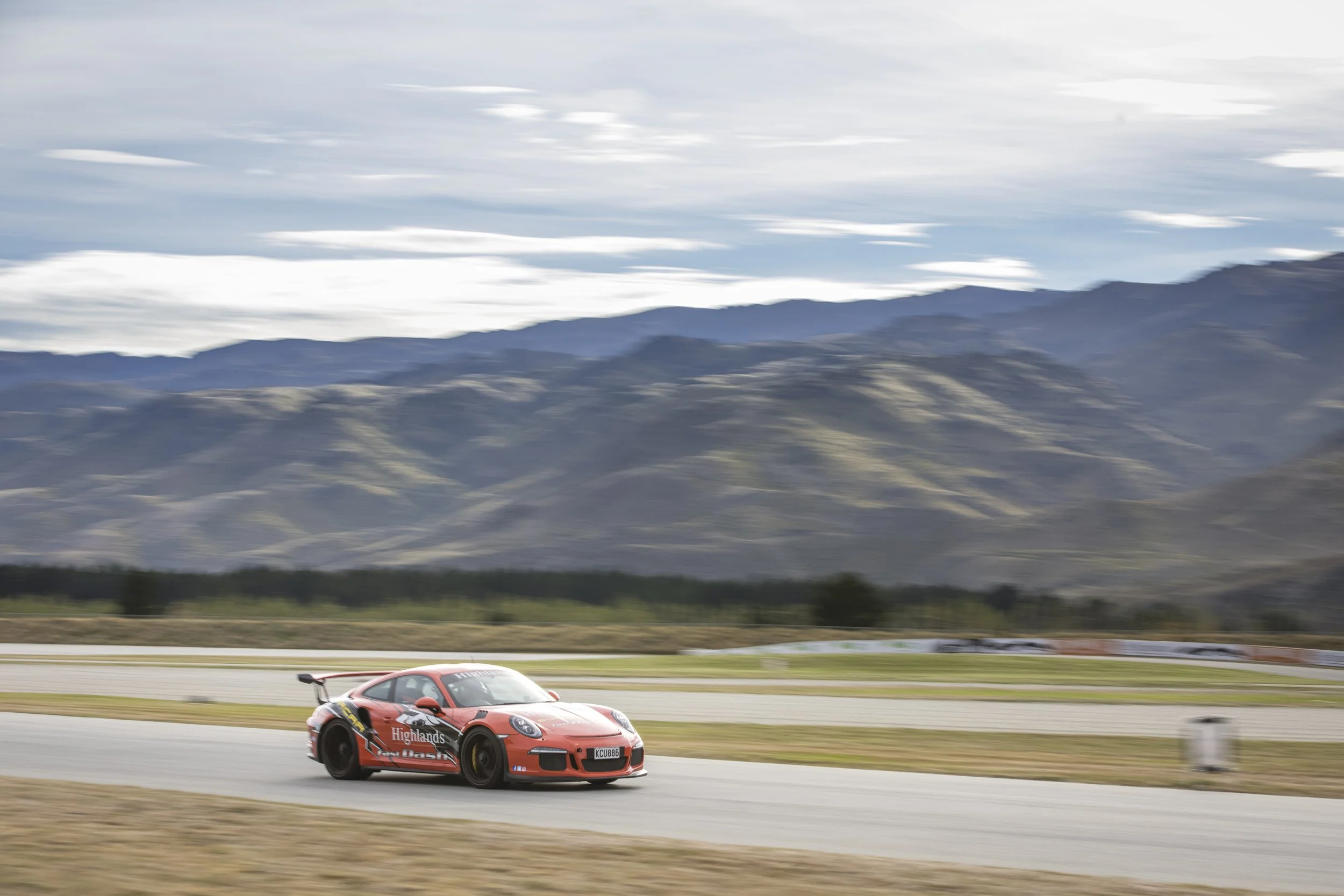 A red sports car racing on a track with mountains and cloudy sky in the background.