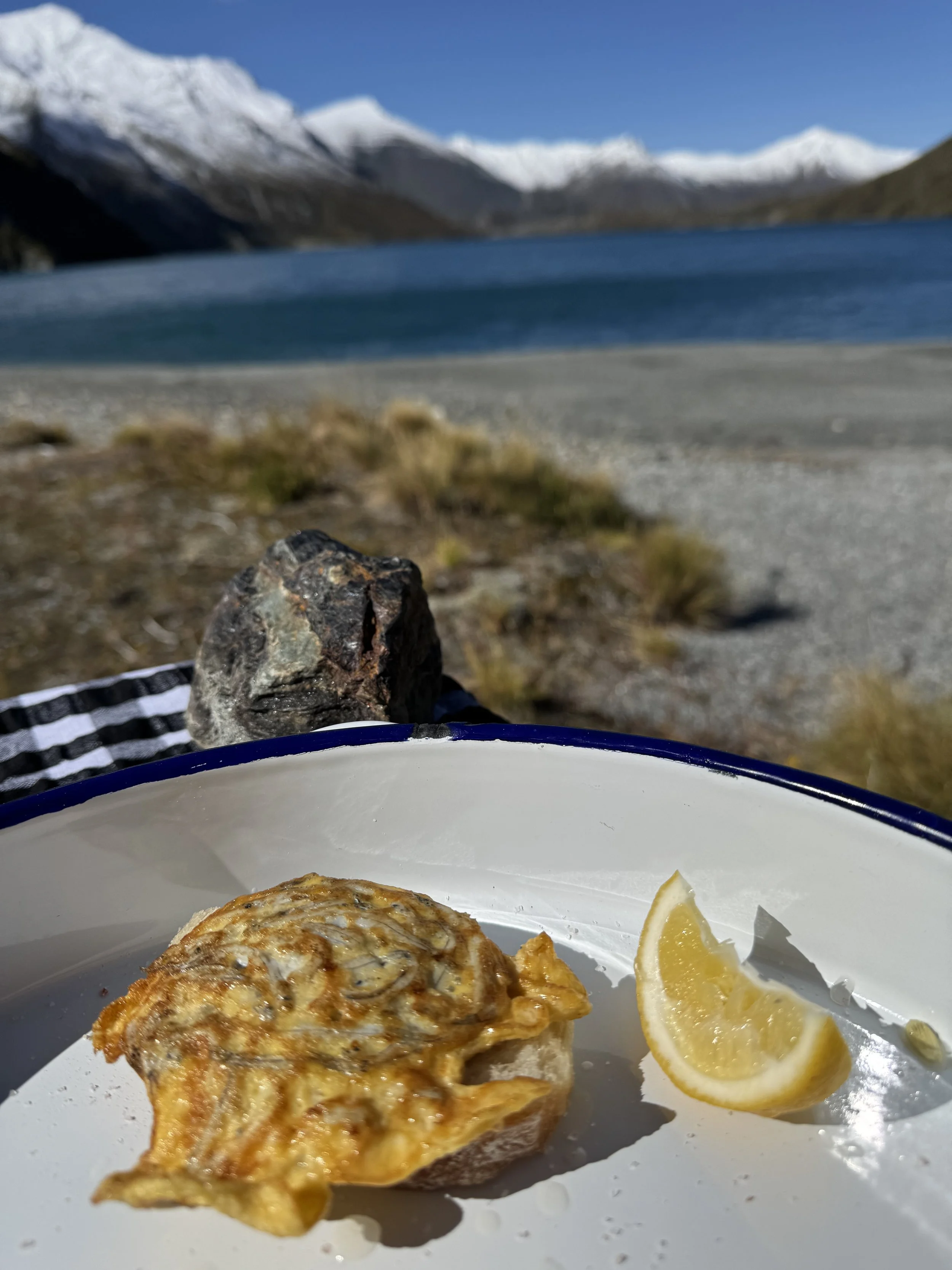A plate with a piece of baked bread topped with melted cheese and herbs, and a lemon wedge, set outdoors near a rocky shoreline with mountains and a blue lake in the background.