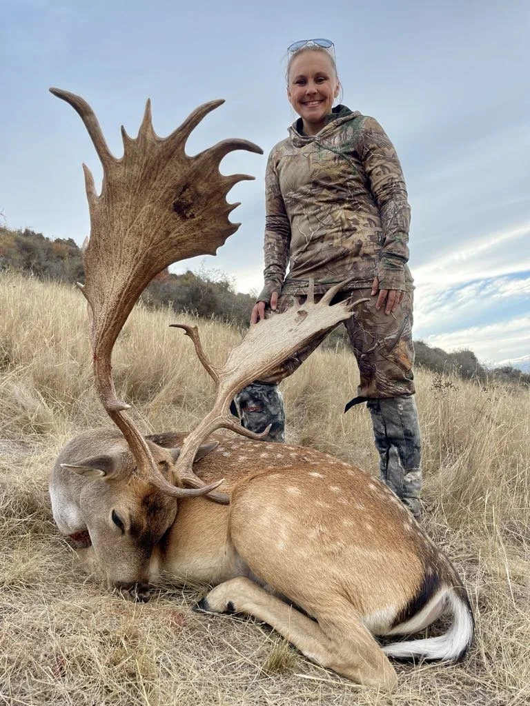 A woman in camouflage clothing standing outdoors on a grassy field, posing with a large dead stag with impressive antlers, which is lying on the ground.