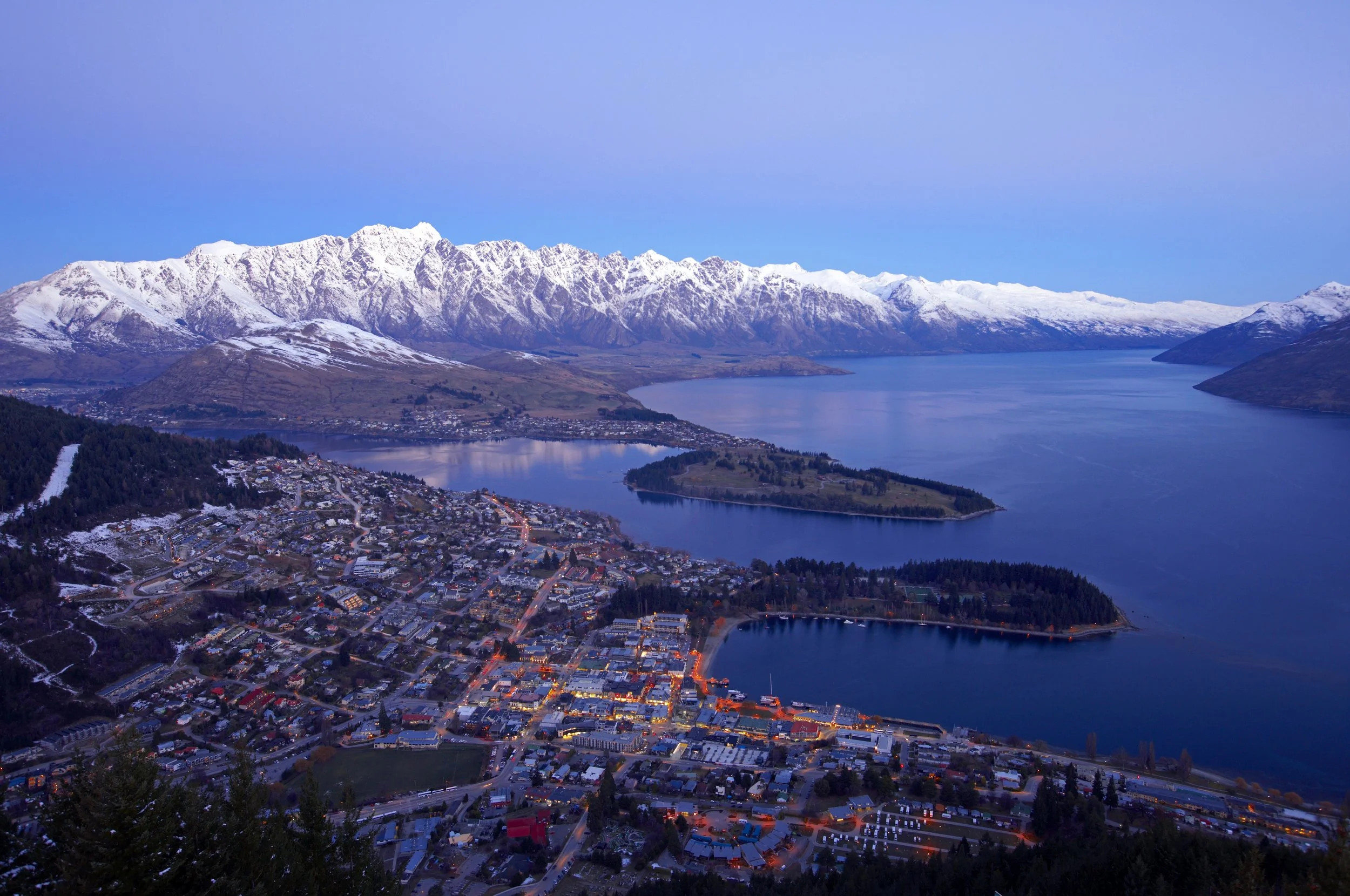 Aerial view of a town with city lights, nestled by a lake, with snow-capped mountains in the background, under a twilight sky.