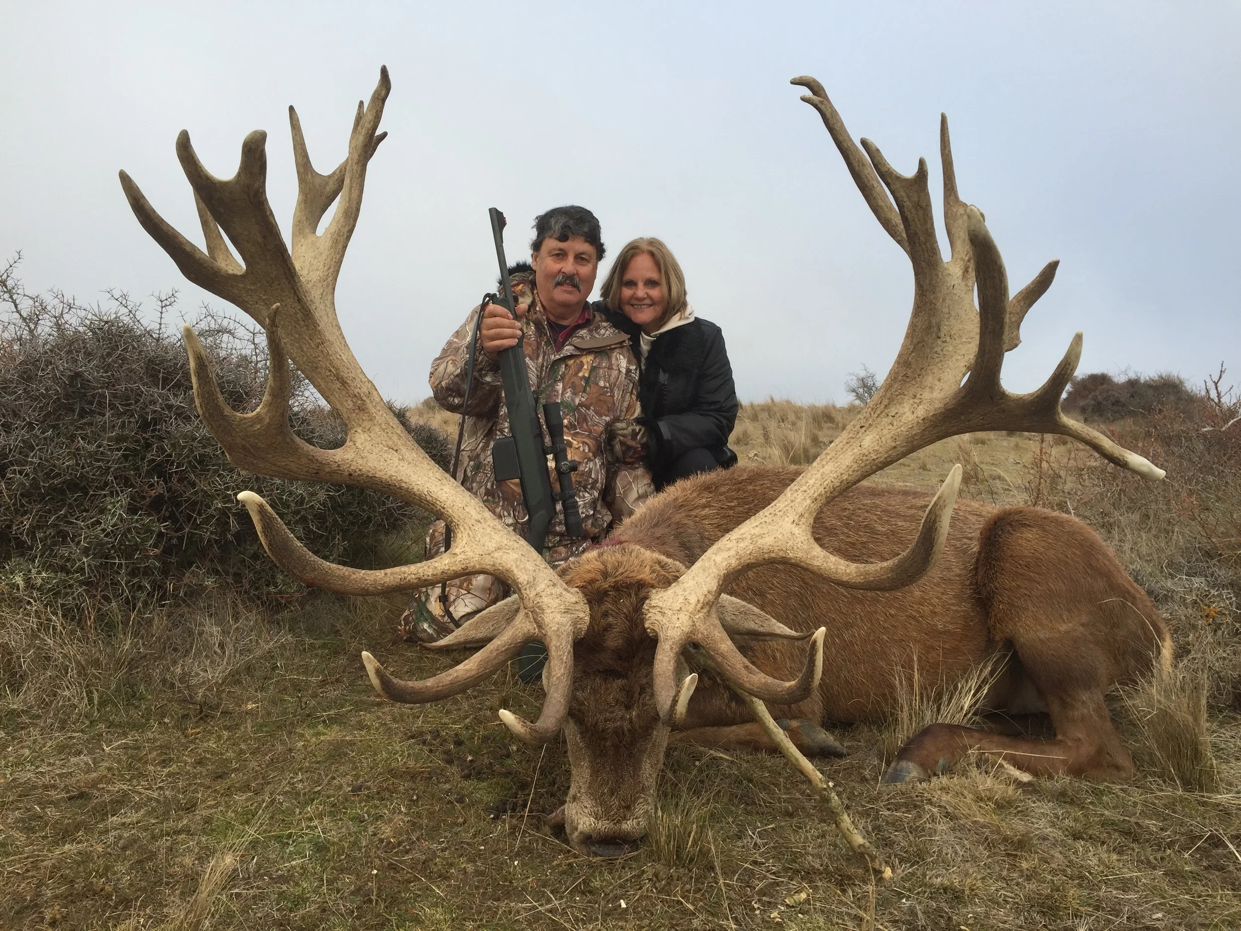 A man and woman standing behind a large, dead stag with impressive antlers, in a grassy, semi-bushy outdoor area.