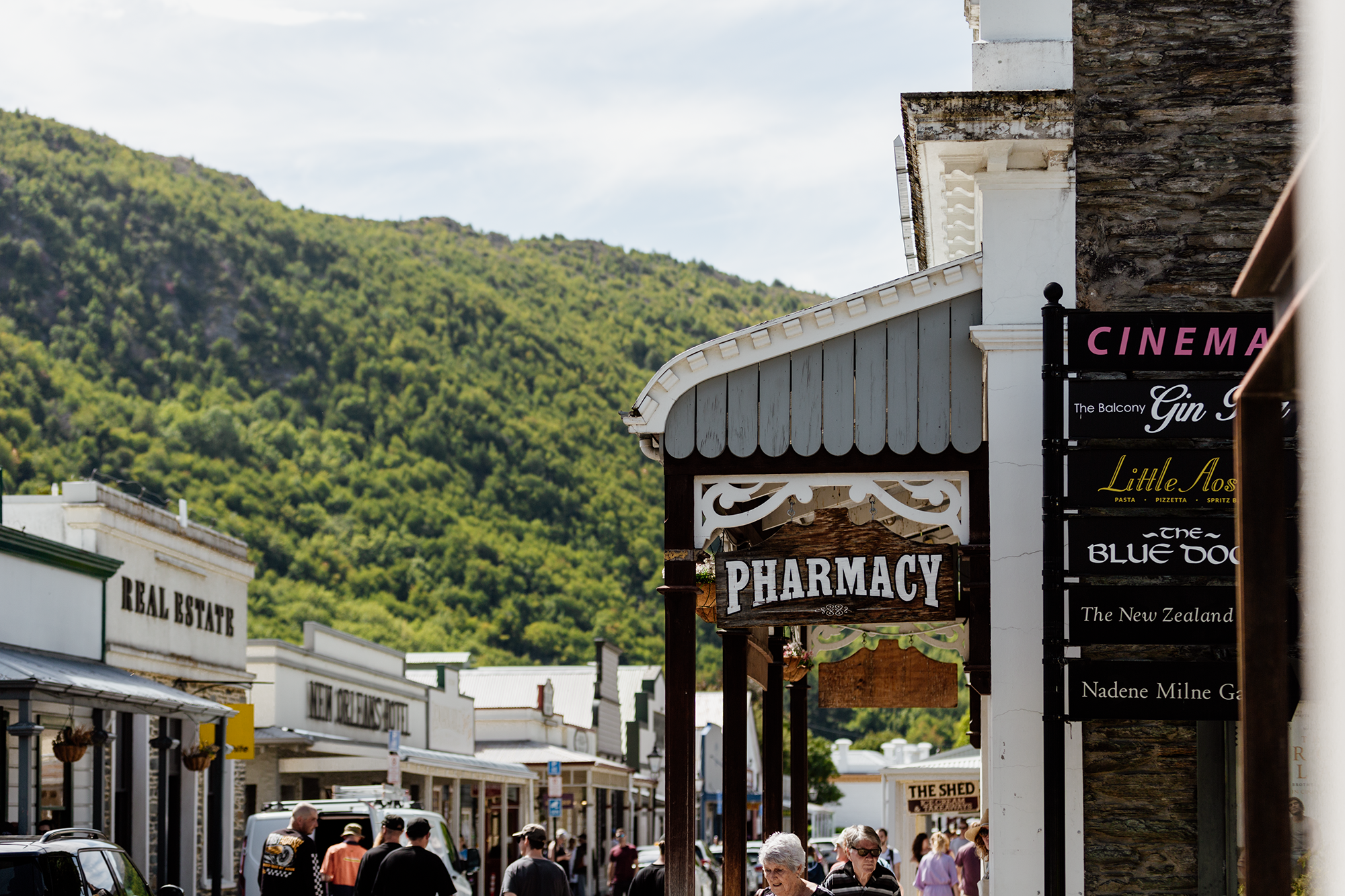 Street scene in a small town with a sign reading 'Pharmacy' in front of a historical building, people walking, and signs for various shops against a green hillside under a partly cloudy sky.