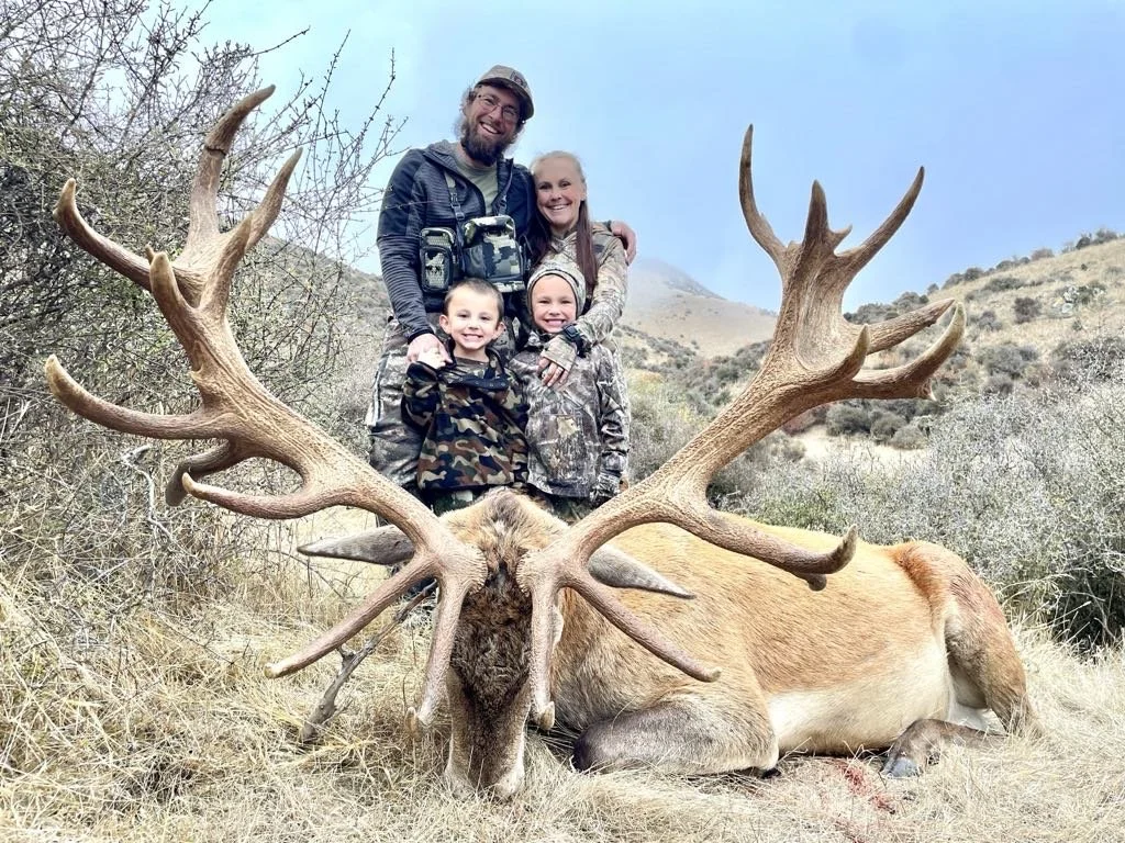 A family of four poses happily behind a large, fallen stag with impressive antlers in a dry, hilly landscape.