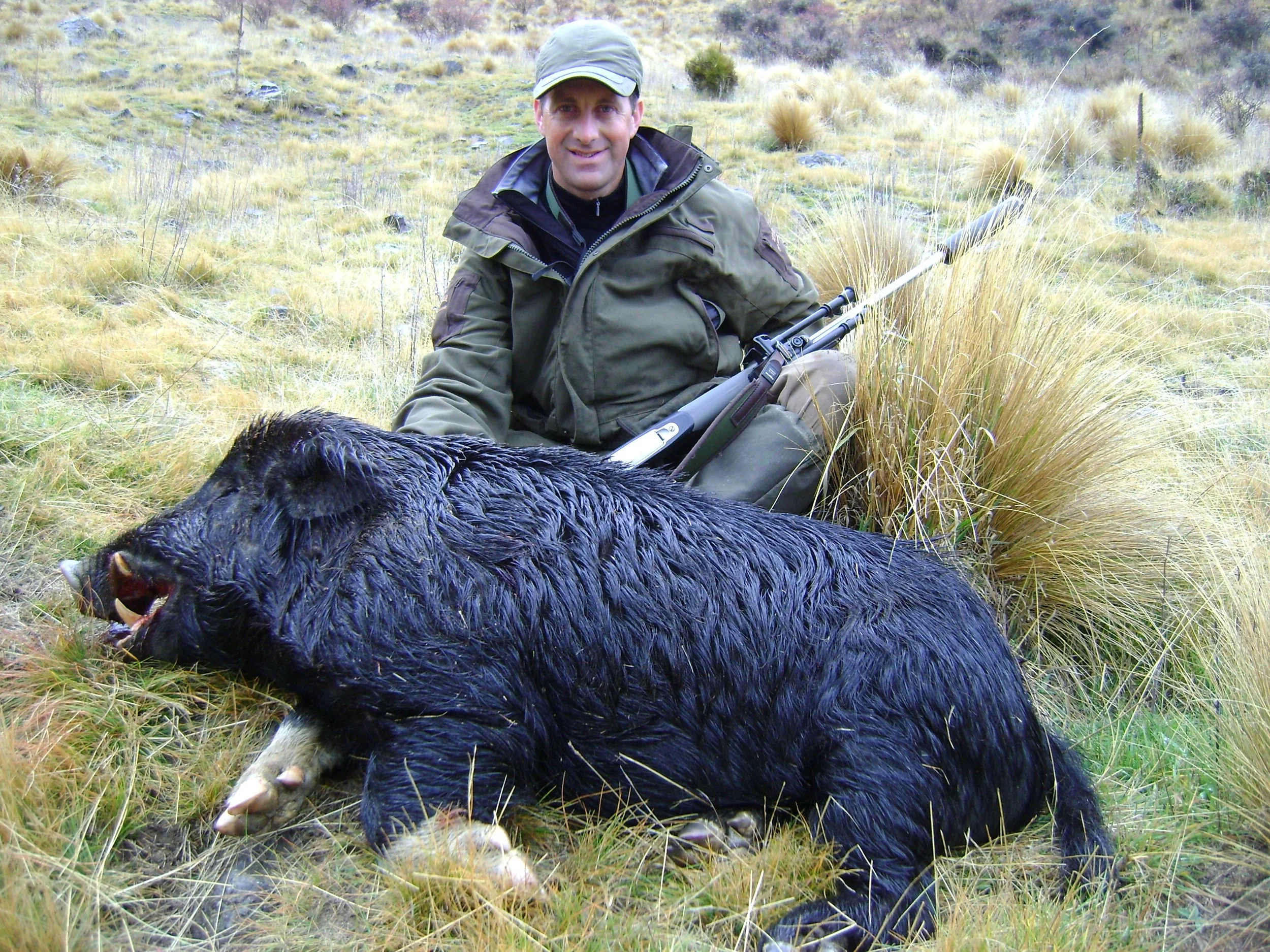 A man sitting on grass in an outdoor setting with a rifle next to a dead black bear.