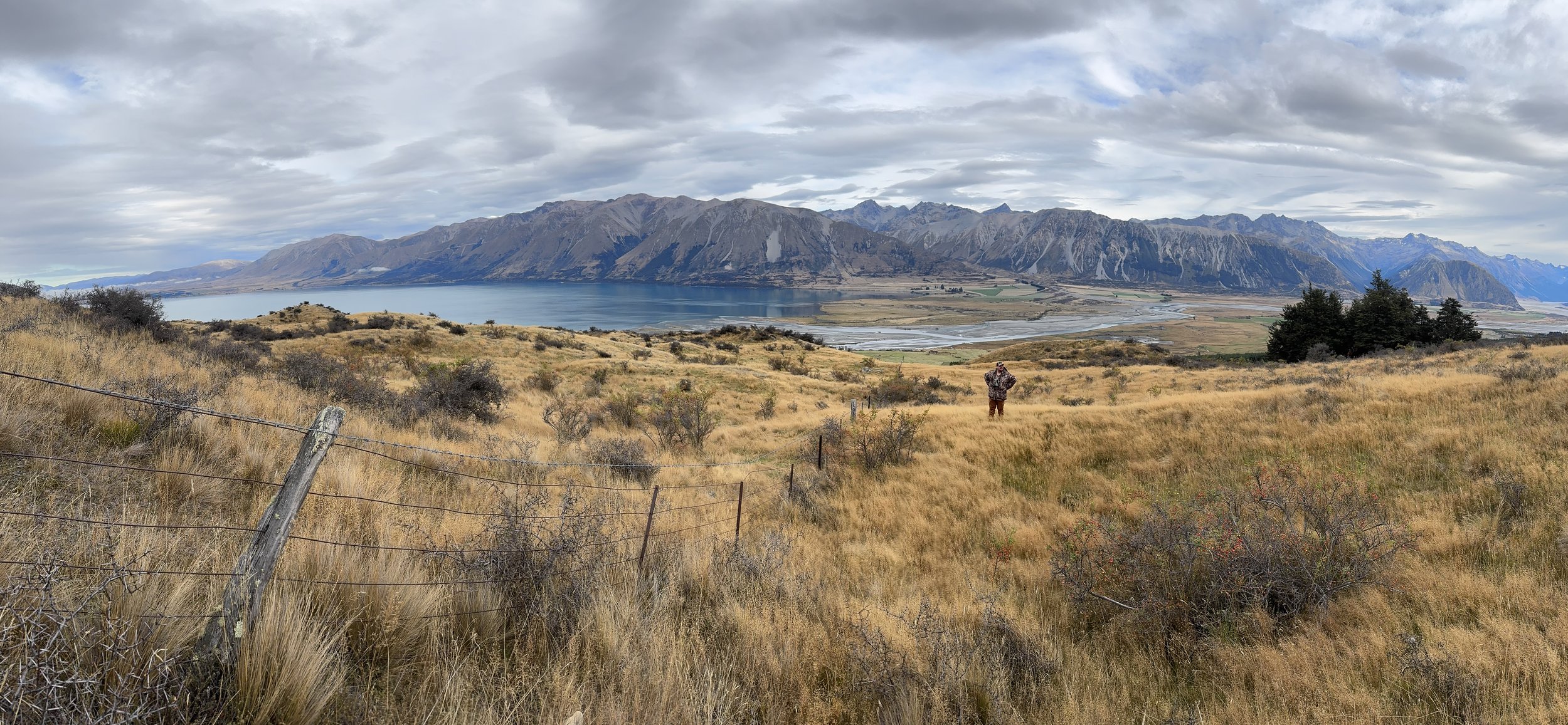 A person in camouflage jacket standing in a grassy field overlooking a lake with mountains in the background.