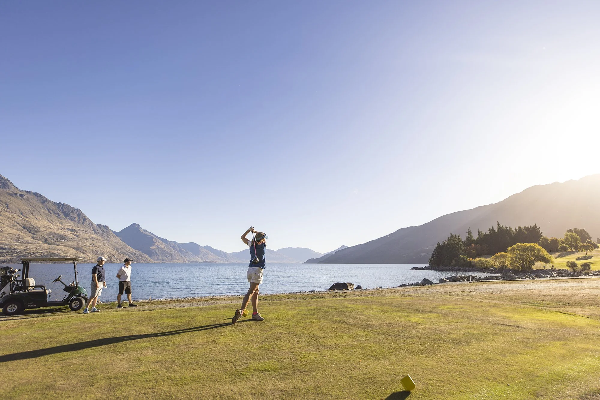 Four people on a golf course near water with mountains in the background, one person swinging a golf club.