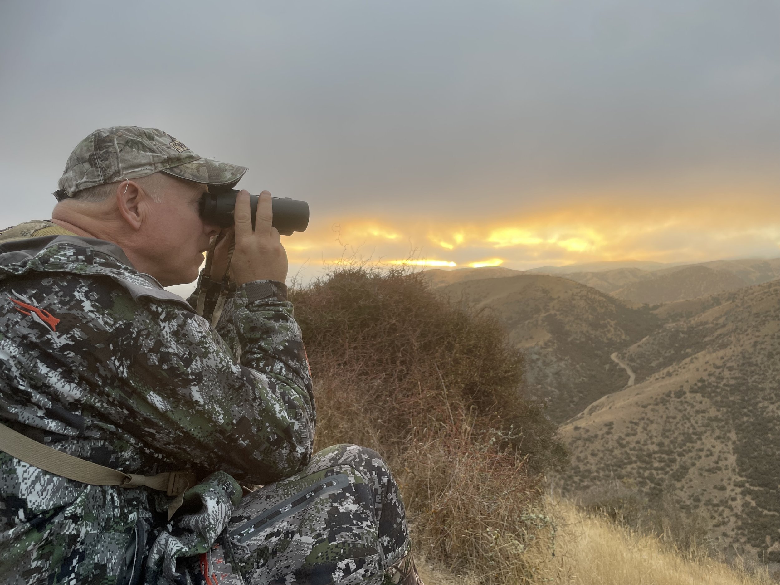 A person in camouflage clothing and a cap using binoculars during sunset in a mountainous area.