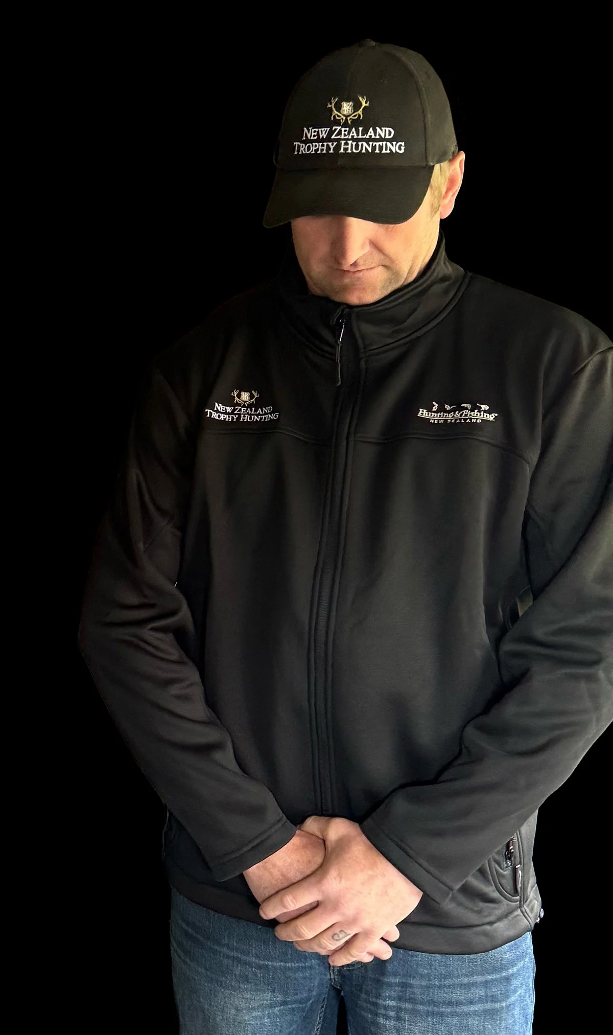 Man wearing a black cap and a black jacket with wildlife and trophy hunting logos against a black background.