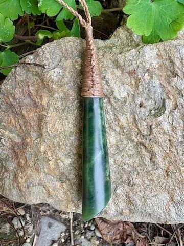 Green stone pendant with a brown braided cord, hanging against a large rock with green leaves in the background.
