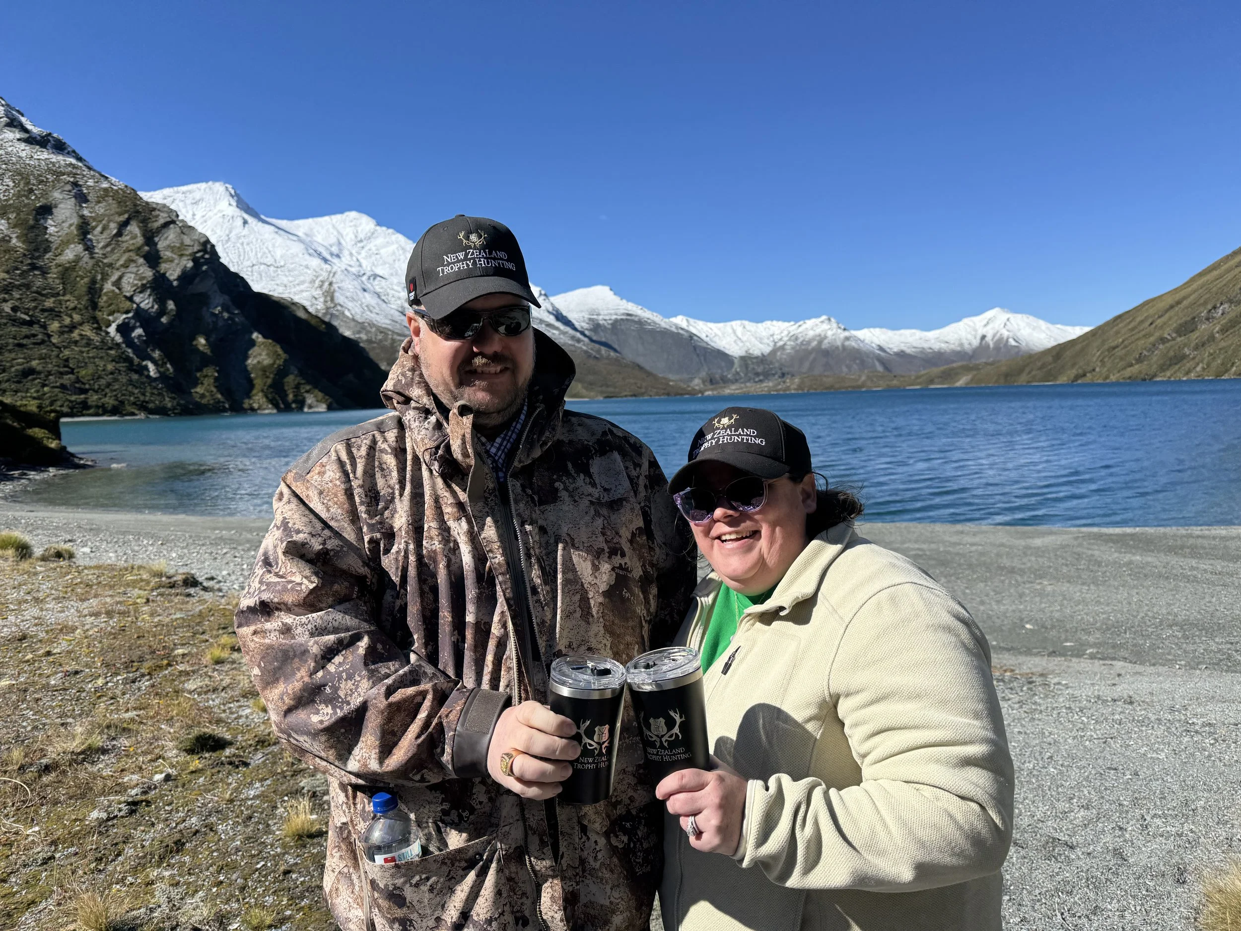 A man and woman smiling and raising drinks in front of a mountain lake with snow-capped peaks in the background, both wearing black hats with 'New Zealand Trophy Hunting' logo.