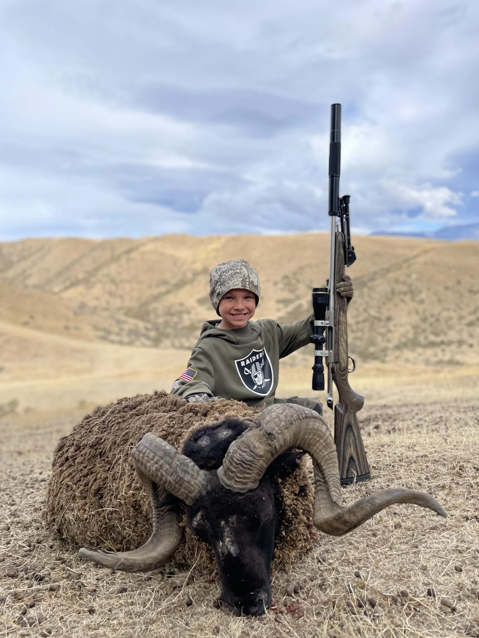 A young boy in camouflage clothing and a Raiders hoodie, sitting on a sheep with large curled horns, holding a hunting rifle, outdoors in a hilly, open landscape with cloudy sky.
