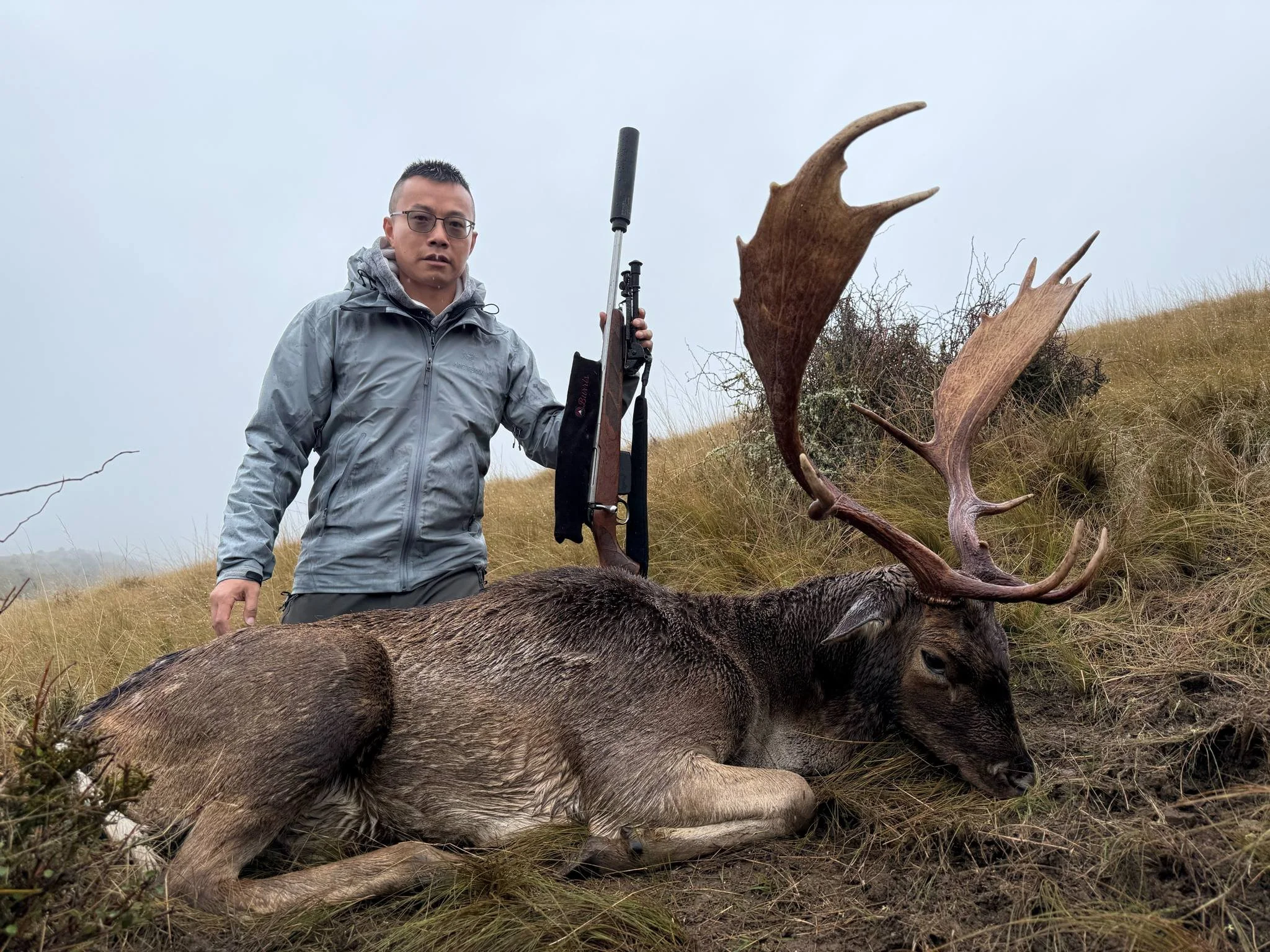 A man in a gray jacket holding a rifle standing next to a large, dead deer with antlers lying on the grass in an outdoor setting with a cloudy sky.
