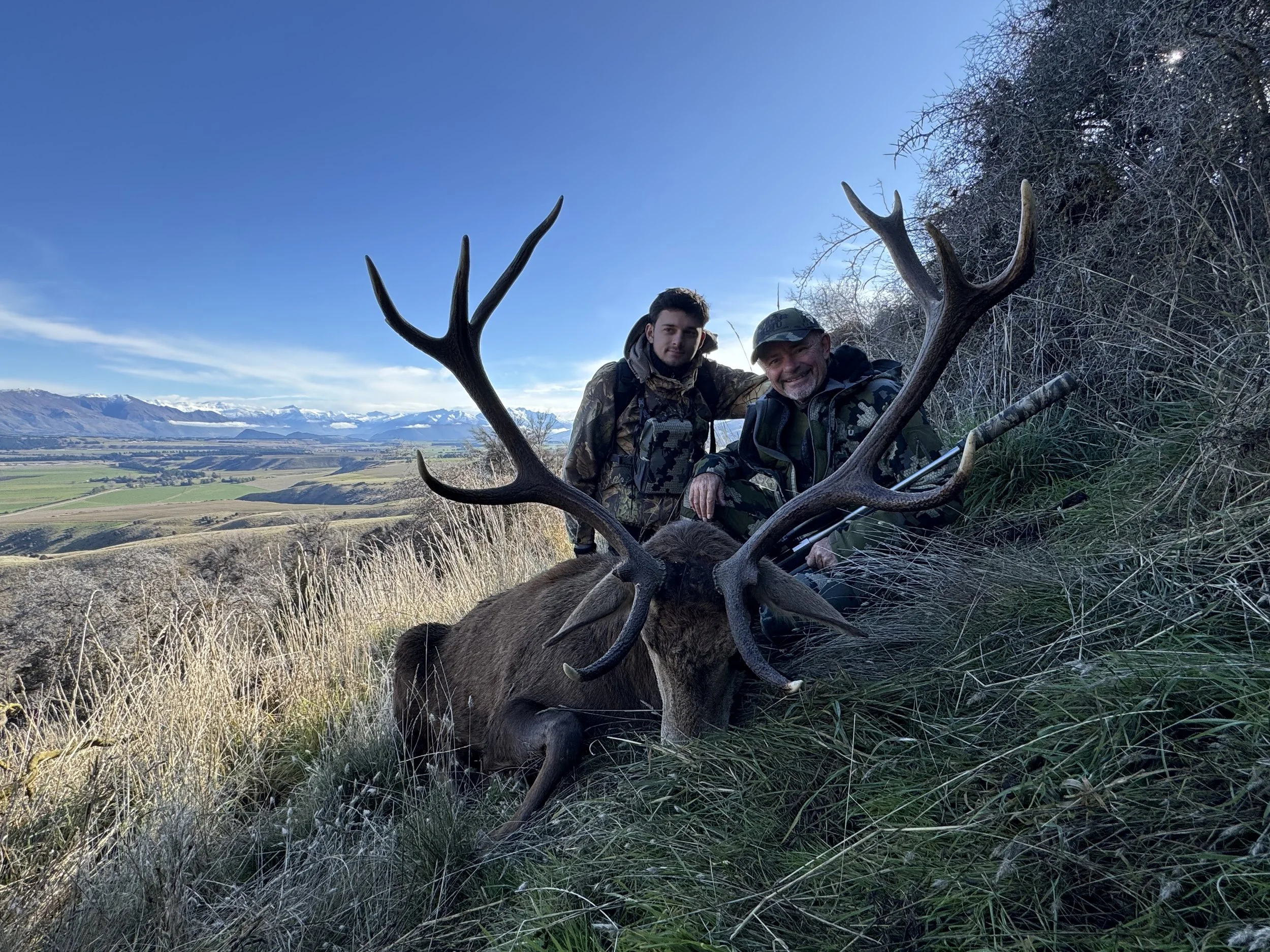 Two hunters pose behind a large black stag with impressive antlers in a hilly landscape with mountains in the distance, under a clear blue sky.
