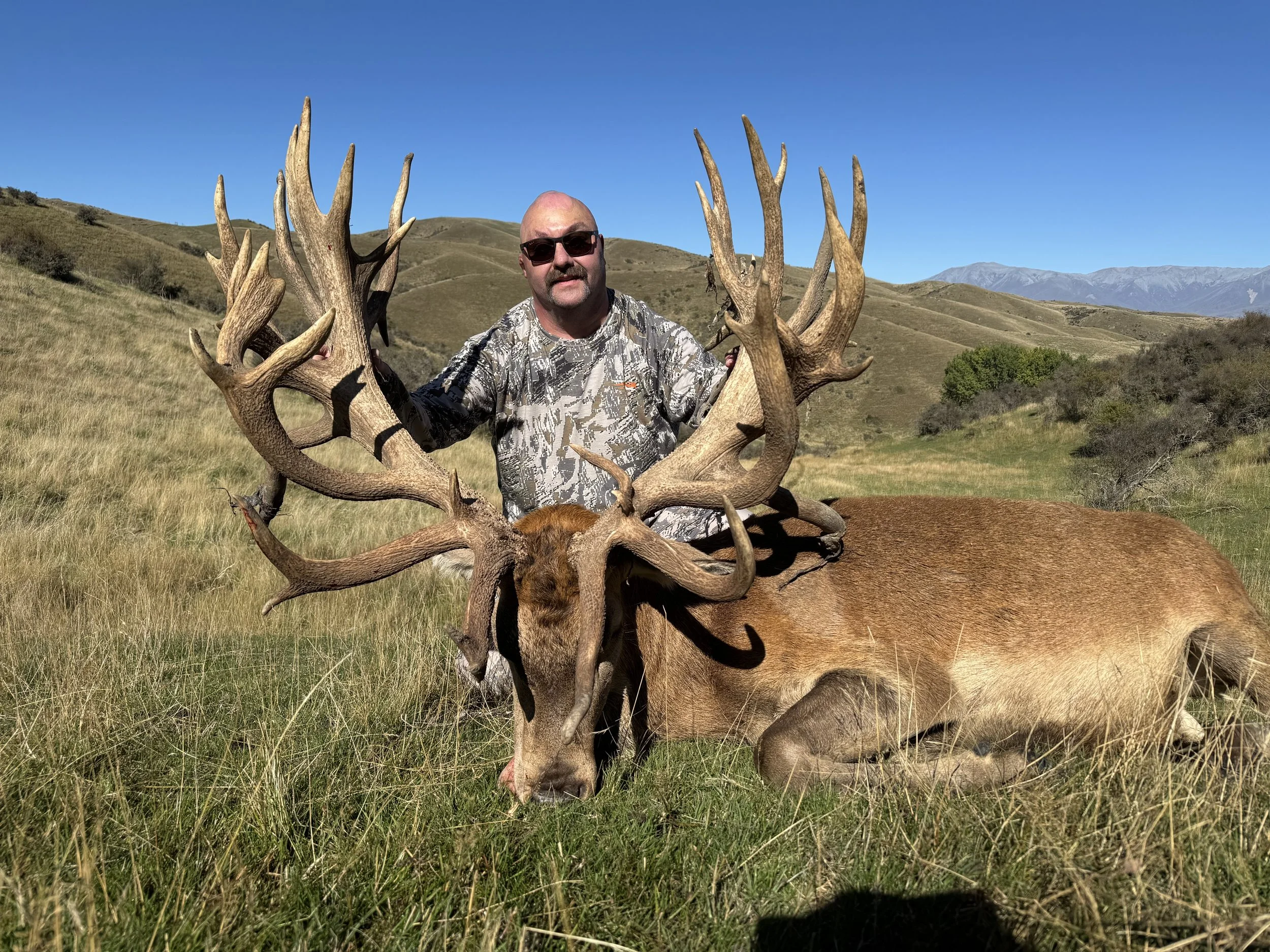 A man in camouflage clothing and sunglasses kneels next to a large, dead elk with impressive antlers in a grassy field with rolling hills and mountains in the background.