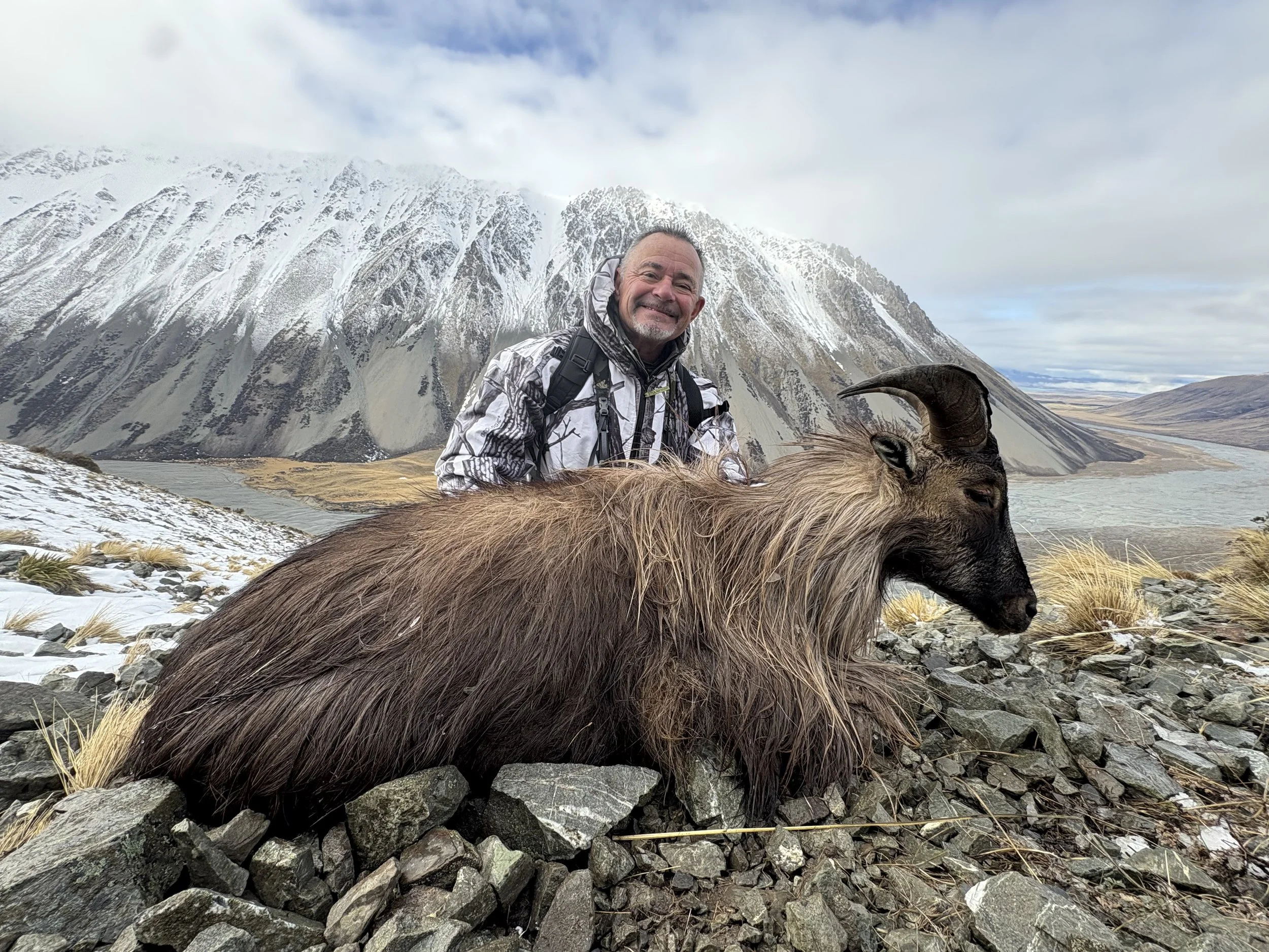 A man in outdoor gear smiling next to a dead goat with large curved horns, set against a mountainous landscape with snow, rocks, and a cloudy sky.