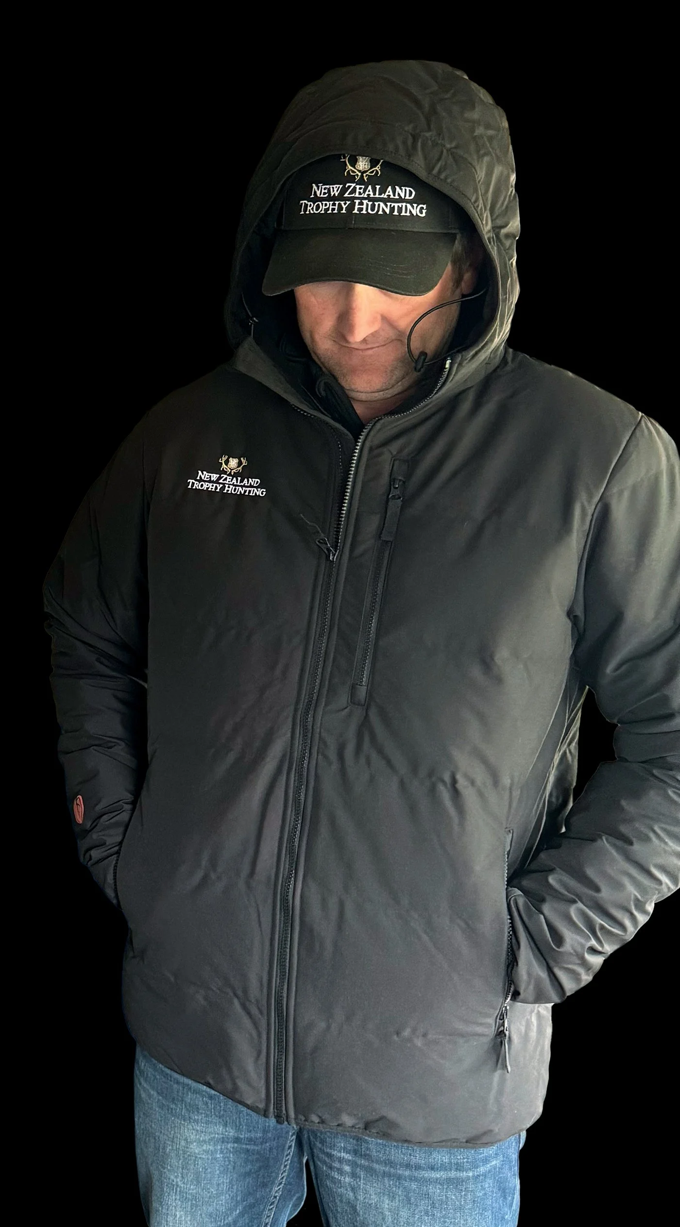 A man wearing a black jacket and a black cap with 'New Zealand Trophy Hunting' written on it, standing with his hands in his pockets, against a black background.