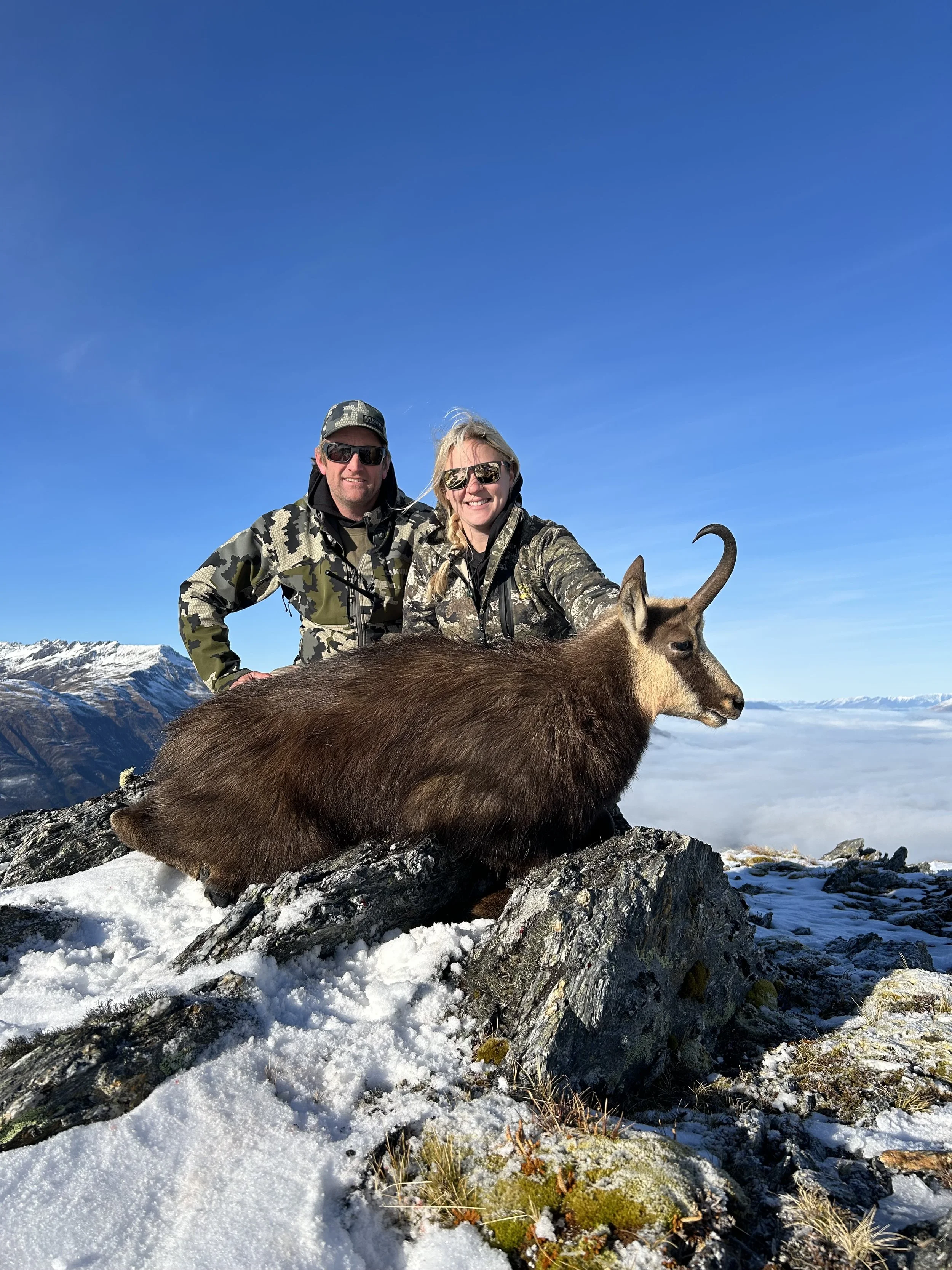 Two people in camouflage clothing standing behind a mountain goat they have hunted, with snow-covered mountains and a clear blue sky in the background.