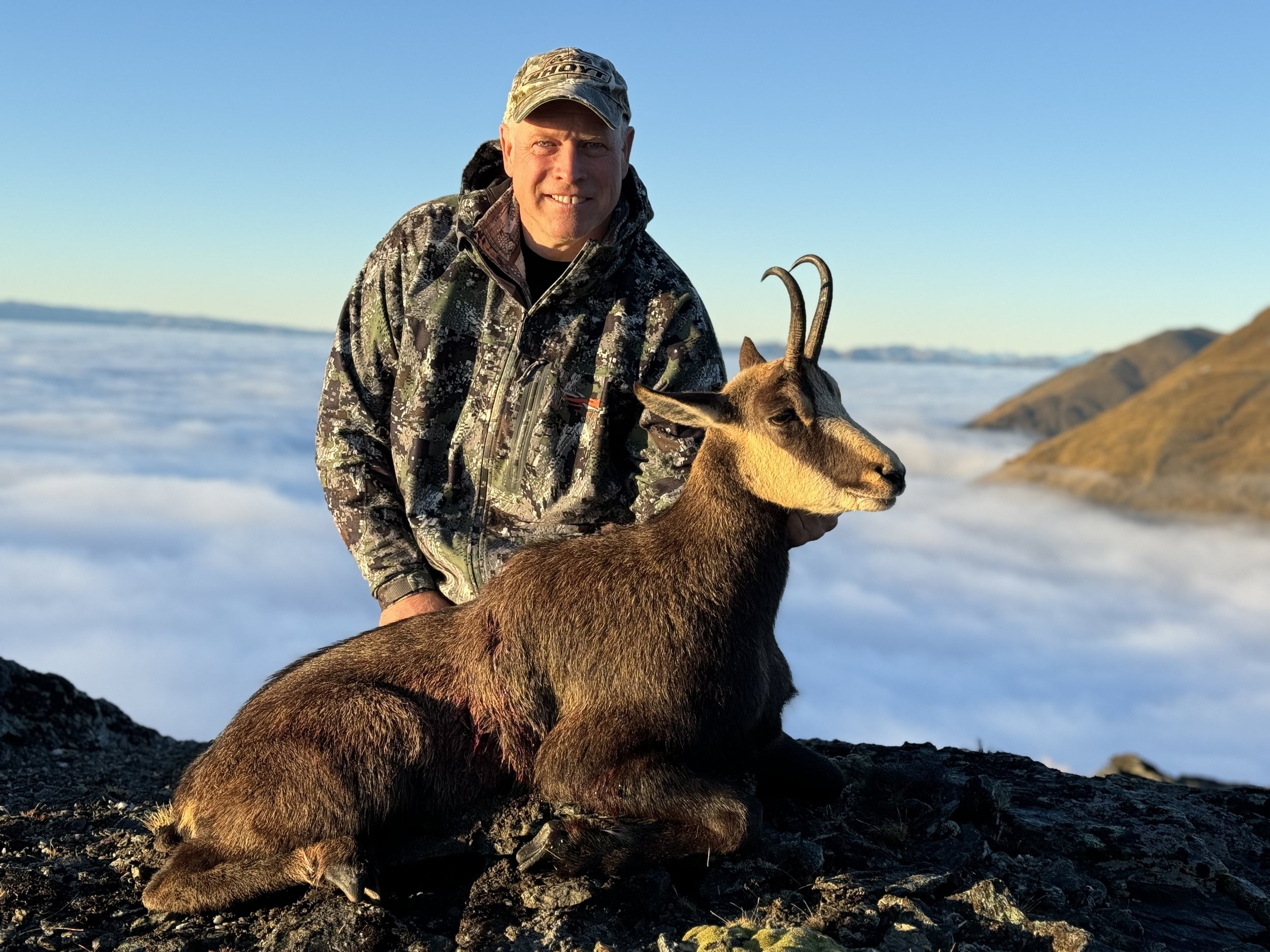 A man in camouflage hunting gear kneels beside a sitting mountain goat with curved horns on rocky terrain, with foggy mountains and a clear blue sky in the background.