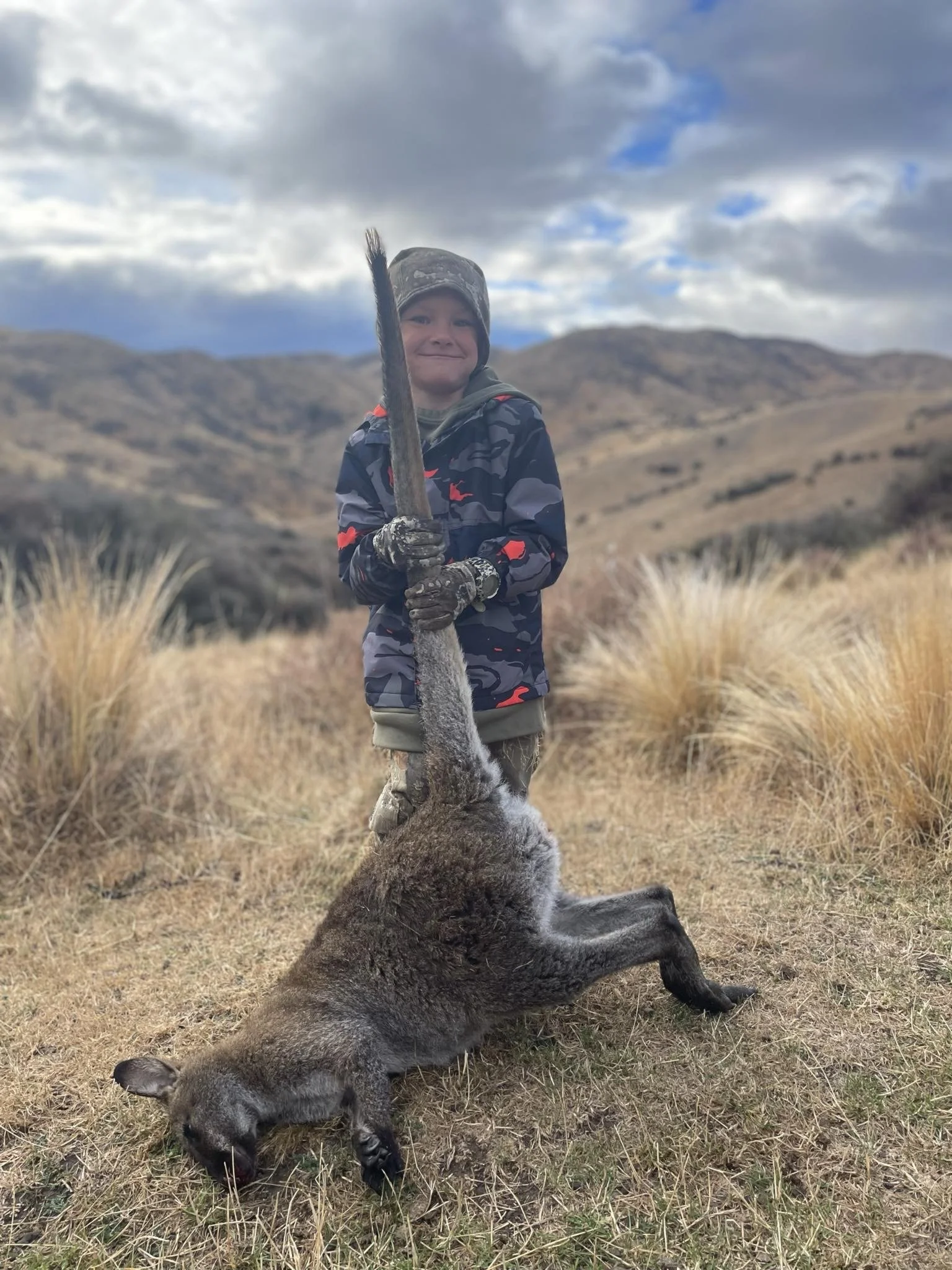 A boy in outdoor camouflage clothing, holding a large hunted animal by the tail, standing in dry grass with mountains and cloudy sky in the background.