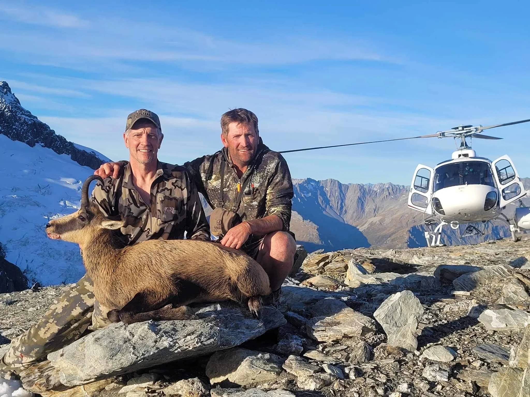 Two men in camouflage clothing pose with a goat on a rocky mountain ridge. A helicopter is flying overhead against a backdrop of snow-capped mountains and a blue sky.