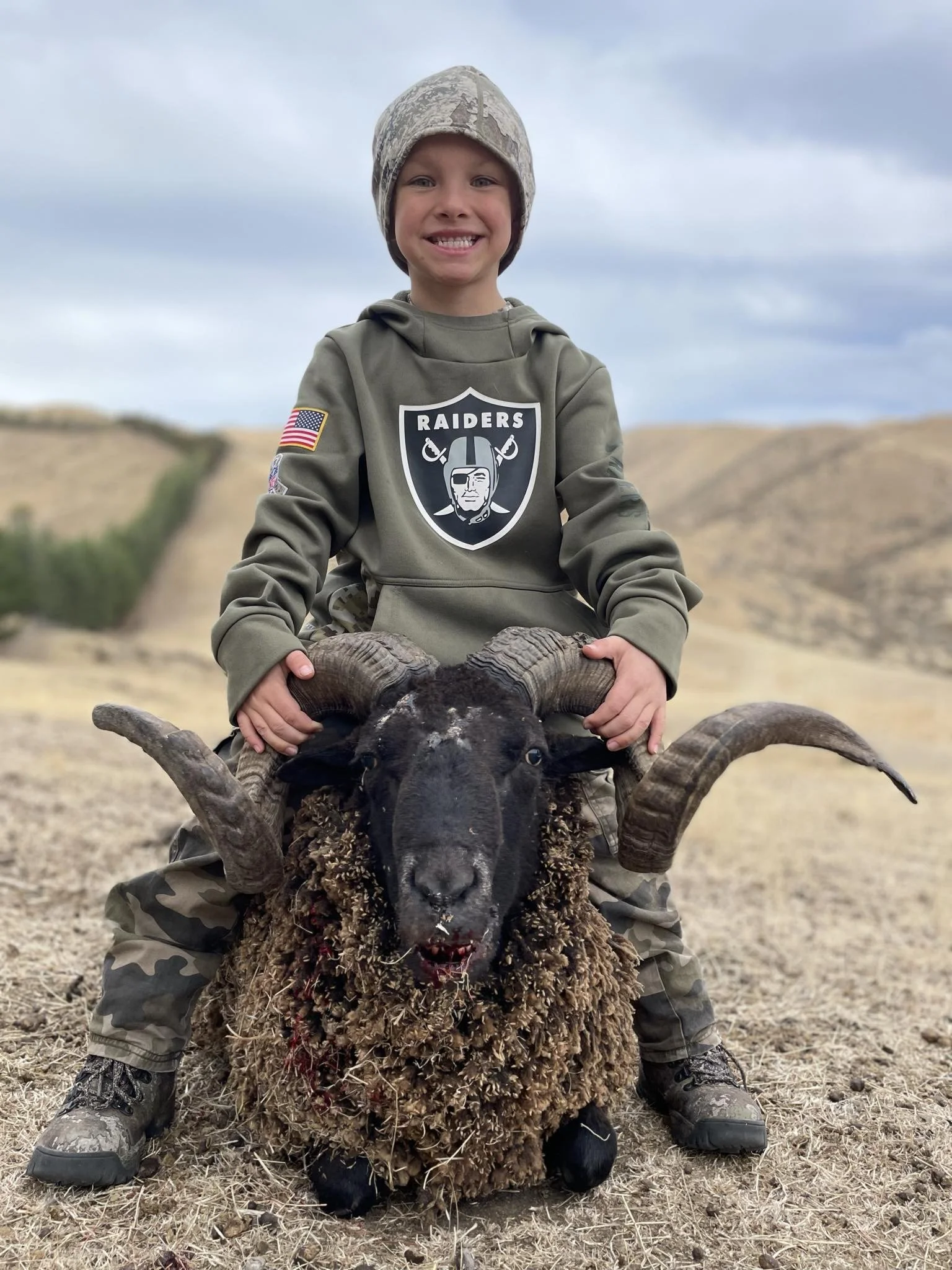 A young boy dressed in camouflage clothing and a Raiders hoodie sitting on a sheep wild in a dry, hilly landscape under a cloudy sky.