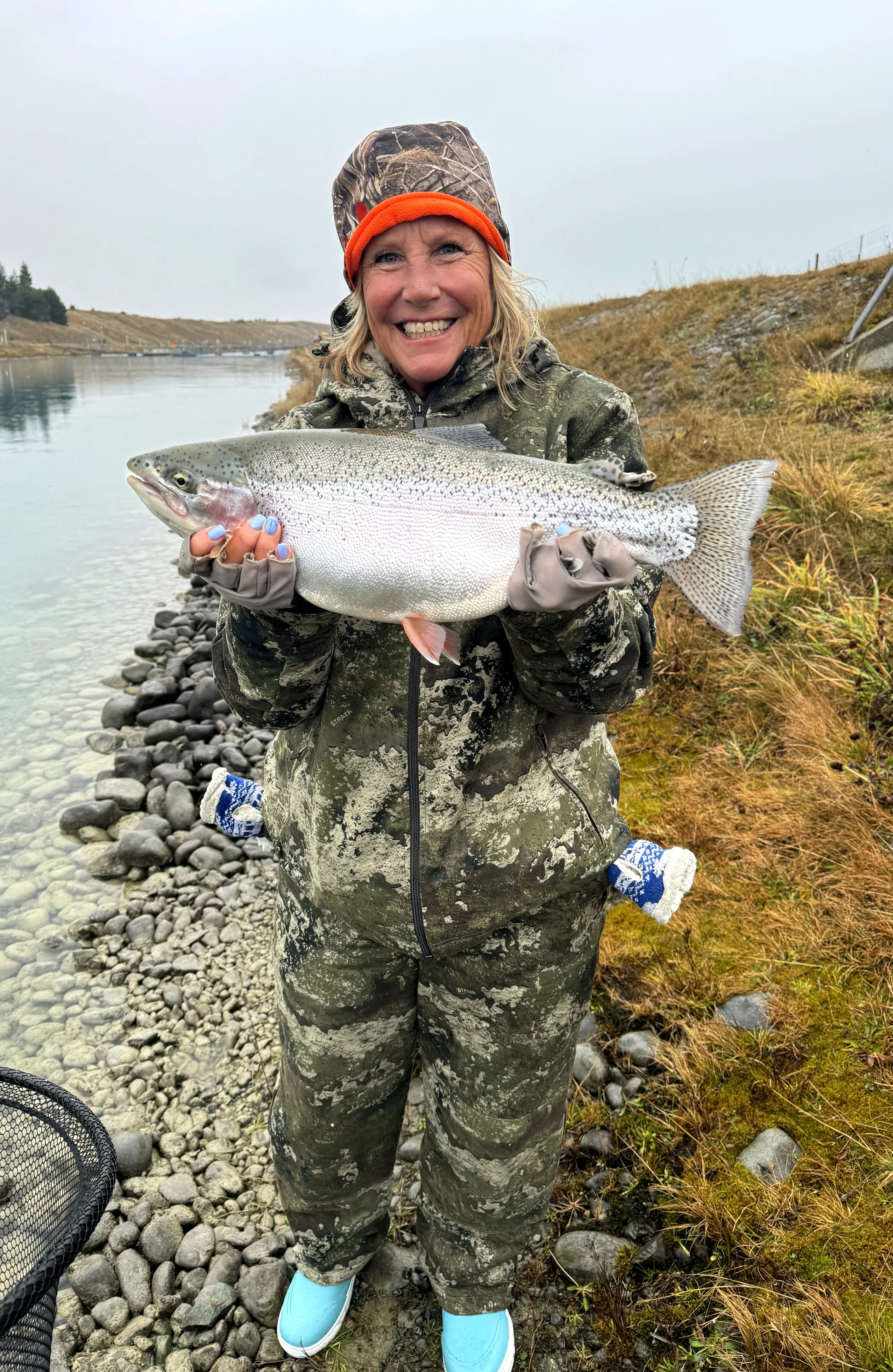 Woman in camouflage clothing and orange headband holding a large trout fish by the water's edge on a rocky shore.
