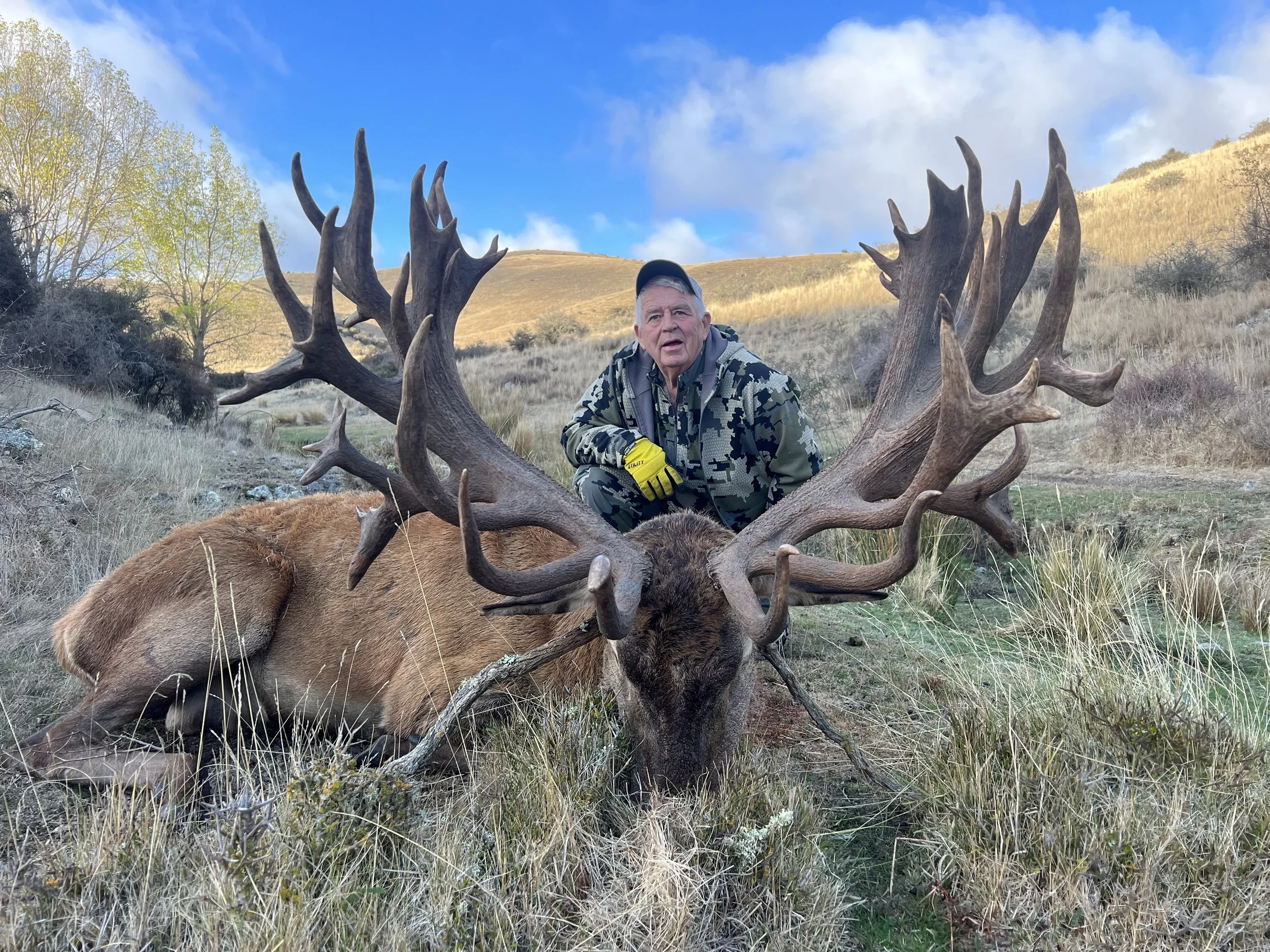 A man in camouflage clothing kneeling next to a large stag with impressive antlers in a grassy field with hills and trees in the background.