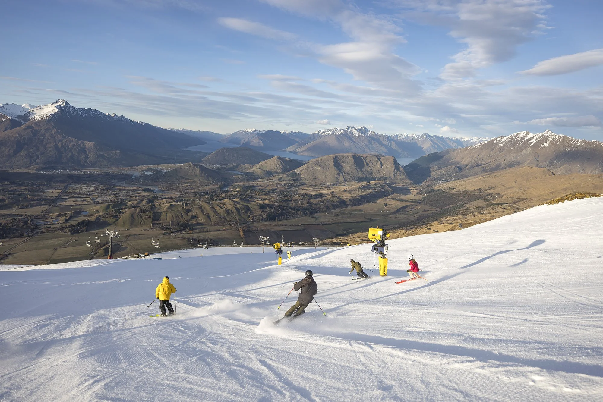 Skier descending a snowy slope with mountains and a lake in the background on a clear day.