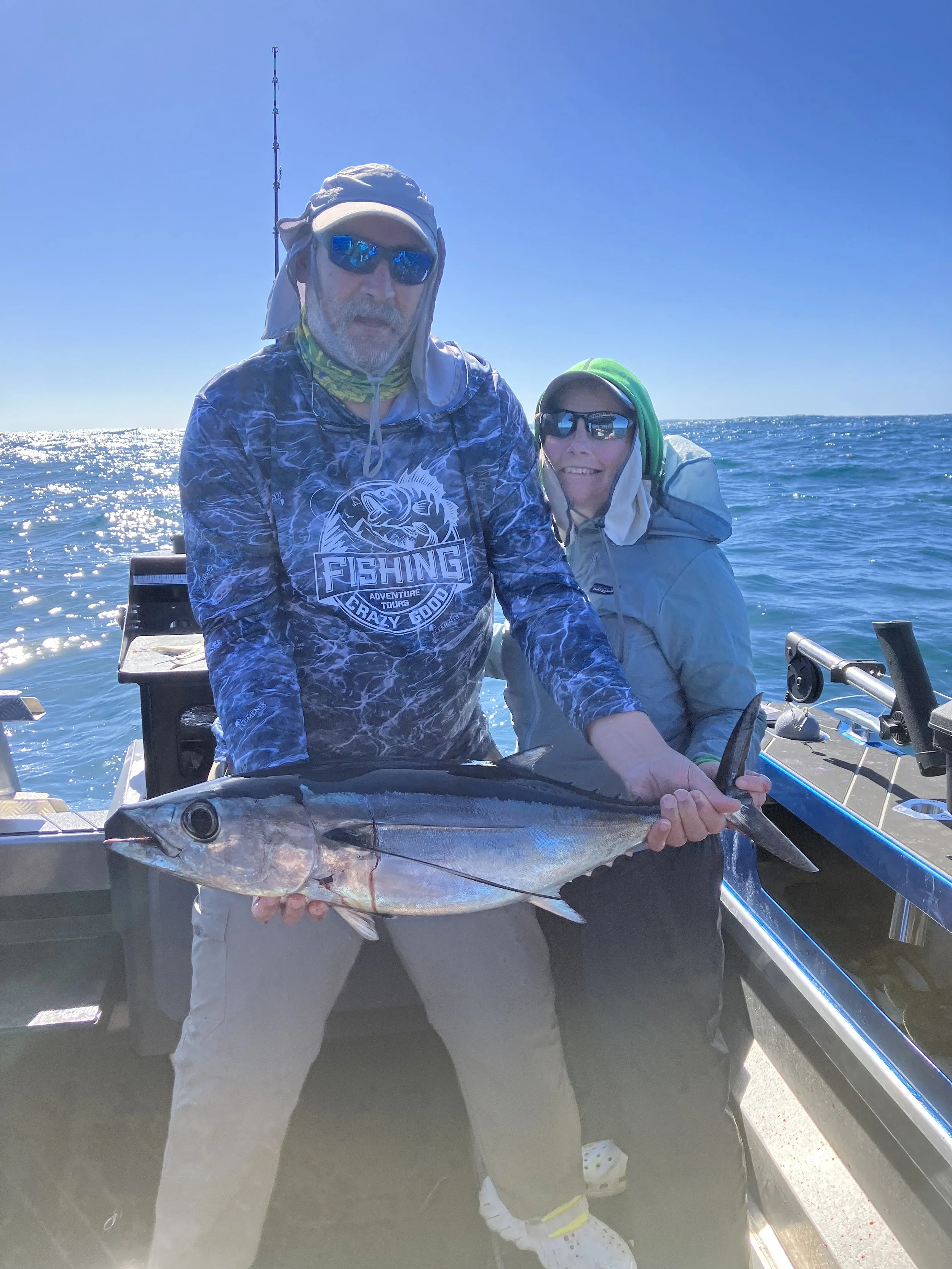 Two people on a boat holding a large fish, with a clear blue sky and ocean in the background.