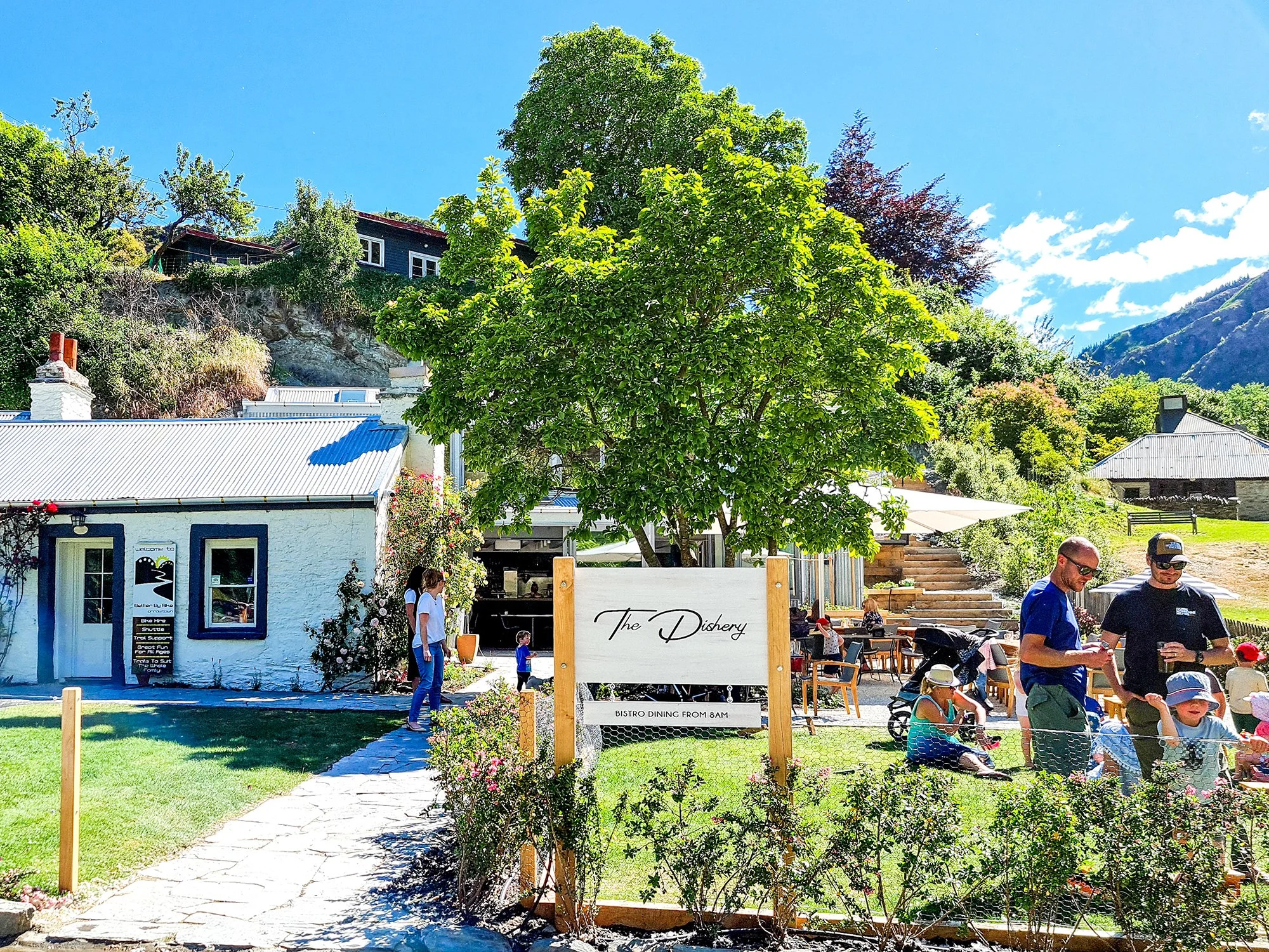A sunny outdoor scene at a cafe called The Dishery with people sitting and walking on a lawn, a large green tree, a white building with black trim, and mountains in the background.