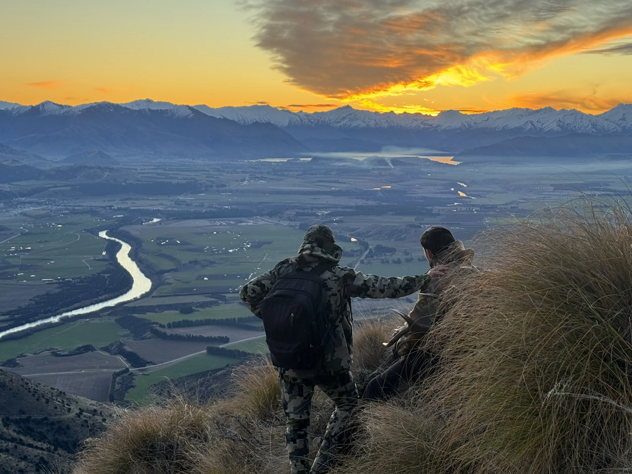 Two men sitting on a grassy hill at sunset, overlooking a river, farmland, mountains, and a colorful sky with orange and yellow clouds.