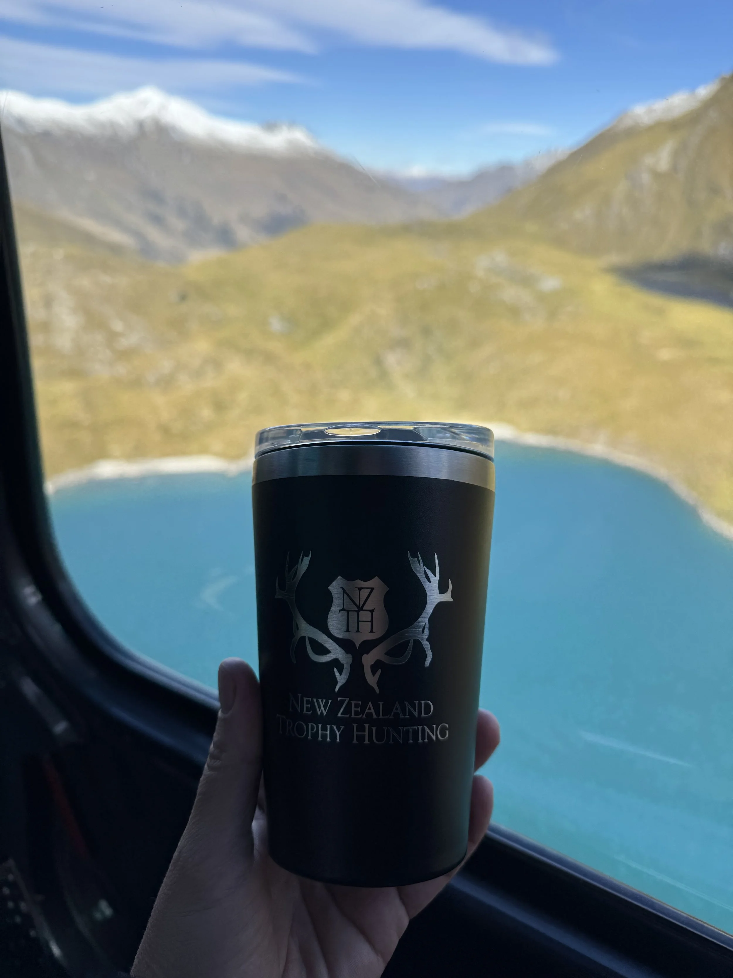 Hand holding a black travel tumbler with 'New Zealand Trophy Hunting' logo, with a mountainous landscape and lake visible through airplane window.
