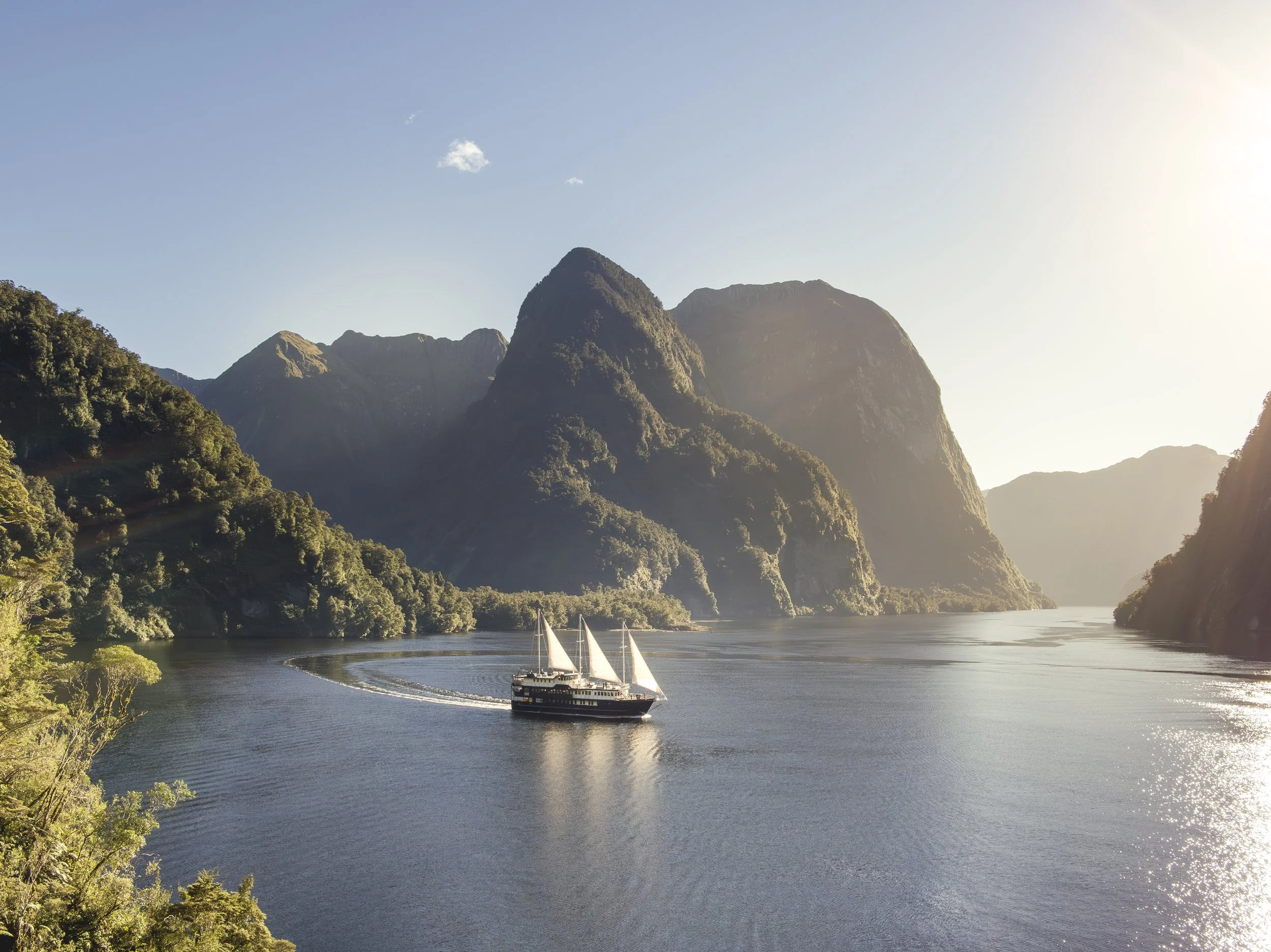 A sailboat with multiple sails sailing on a river surrounded by lush green mountains under a clear sky, with sunlight reflecting on the water.