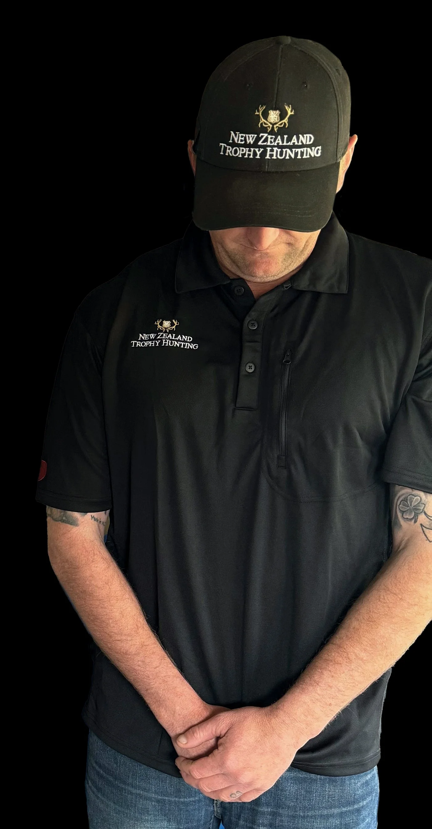 A person wearing a black cap and a black shirt with the logo and text 'New Zealand Trophy Hunting,' standing against a black background, with arms crossed and head slightly down.